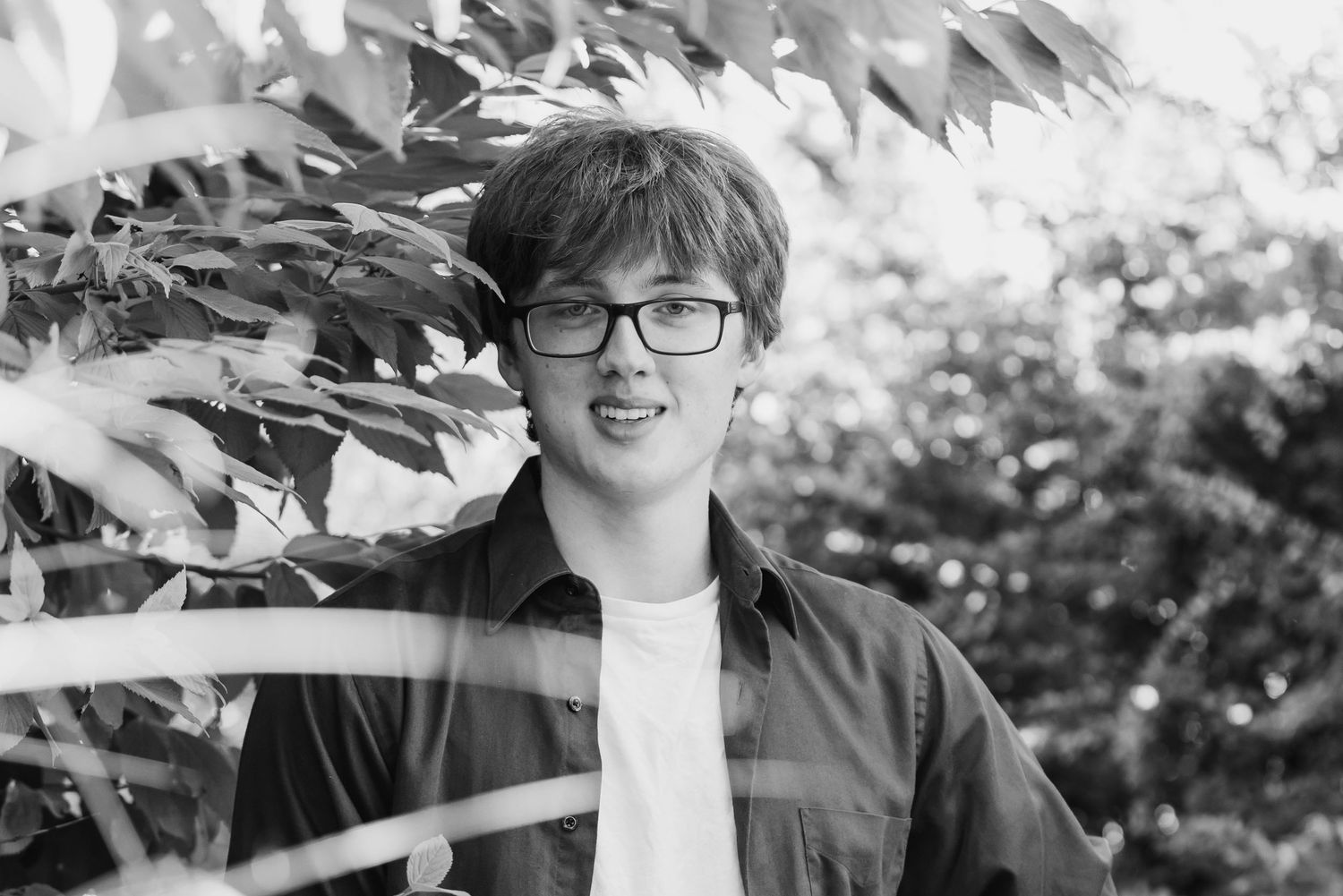 Black and white portrait of someone wearing glasses and smiling outdoors with trees in the background.