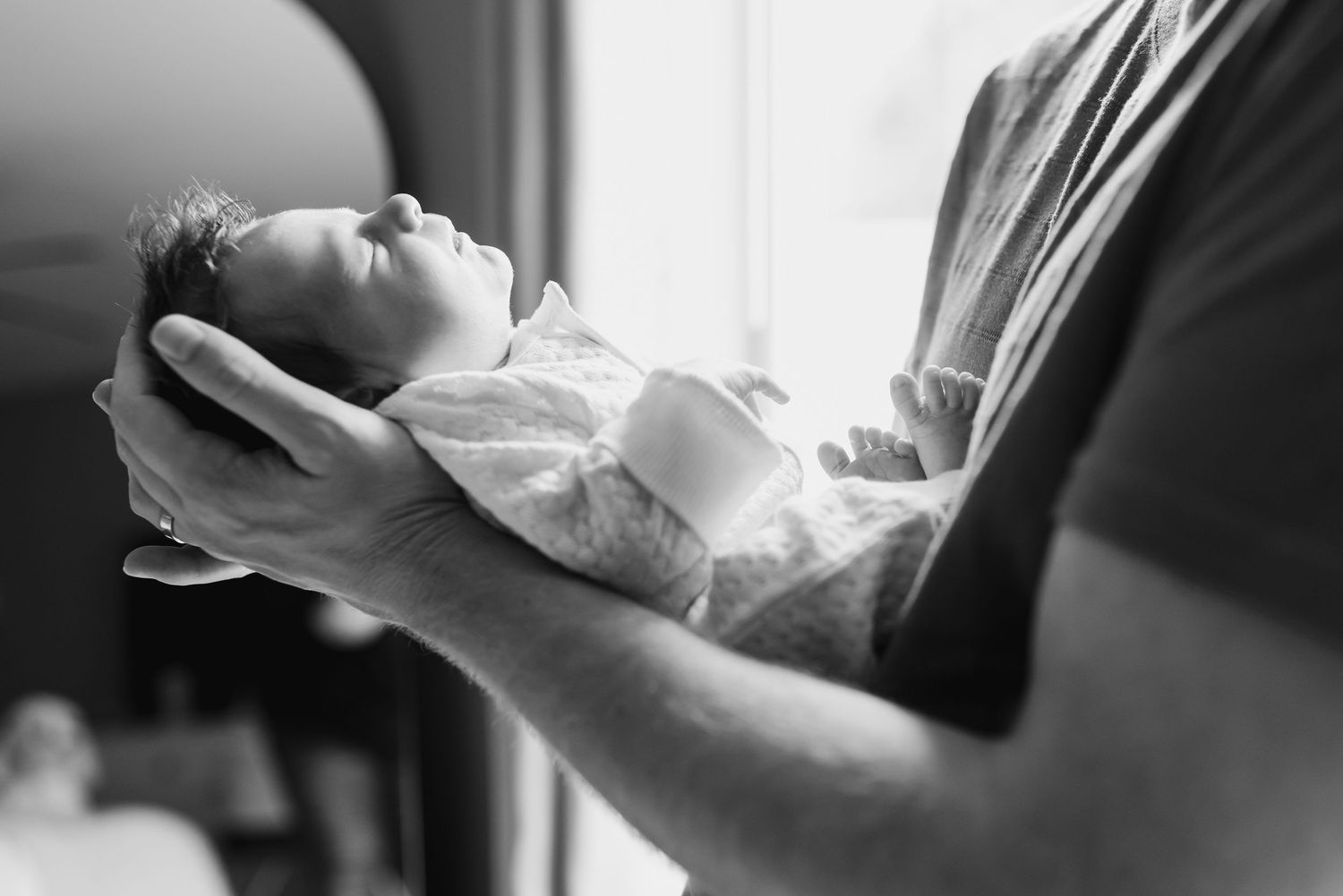 Black and white photo of a new father cradling his newborn son in his hands. The baby is wearing a white onesie and sleeping.