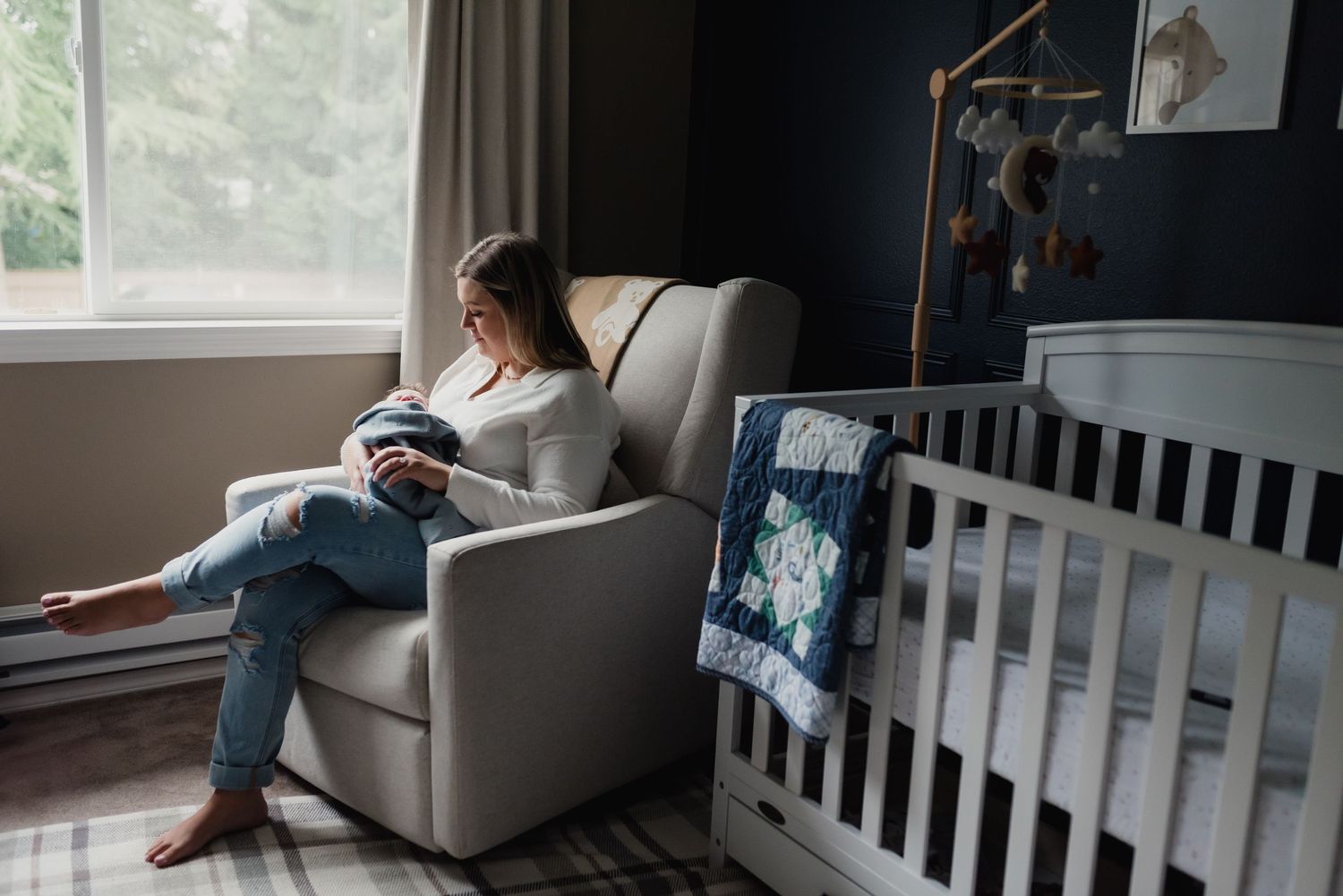 A person sits in a cozy nursery rocking chair next to a window and crib with a blue patterned blanket draped over it.