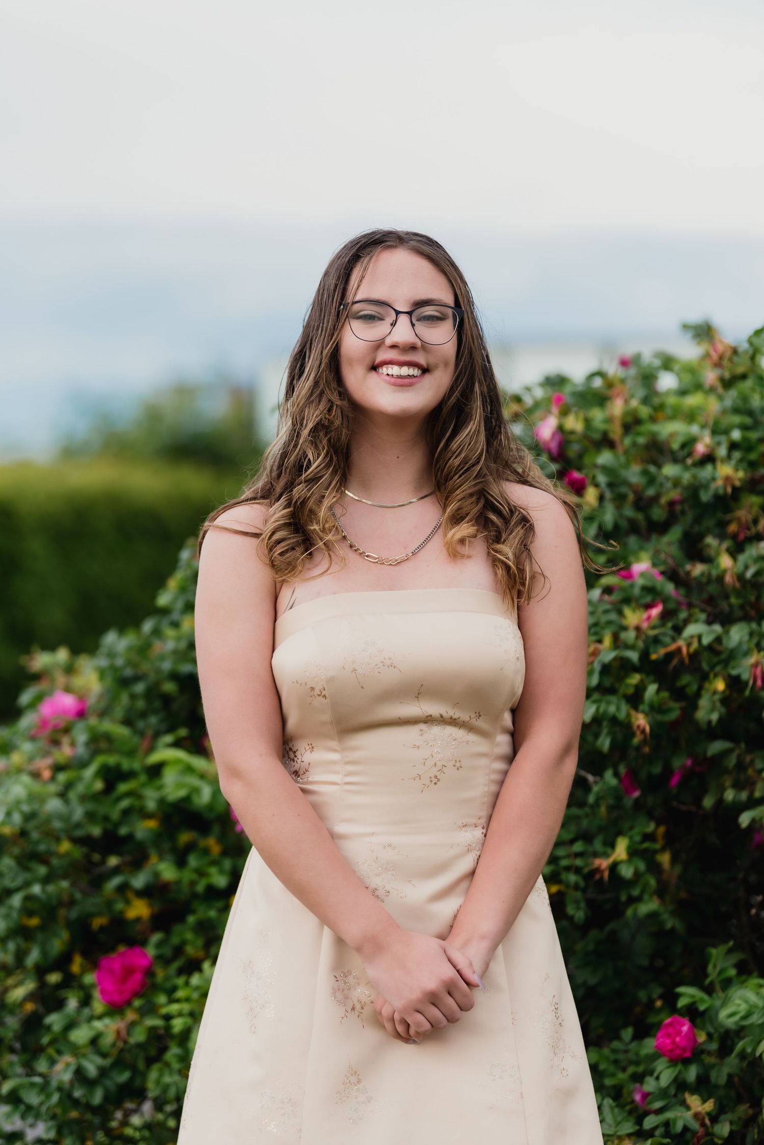 A person in a champagne-colored dress poses against a flowering garden backdrop with pink blooms.