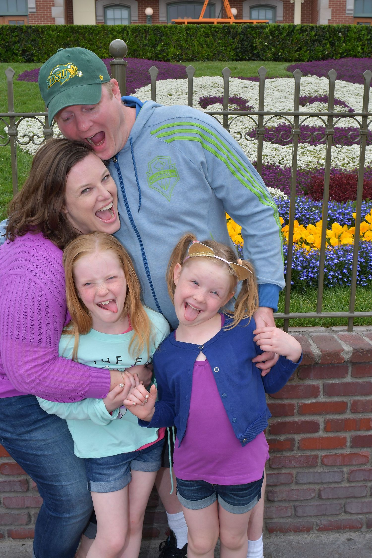 A group poses playfully together against a brick wall with colorful flowers and landscaping in the background.