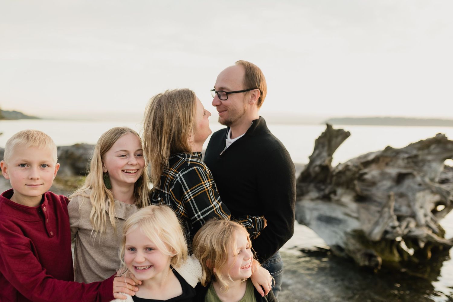 Family of five smiling together on a rocky beach near driftwood in Mukilteo at Lighthouse Park during a casual outdoor portrait session.