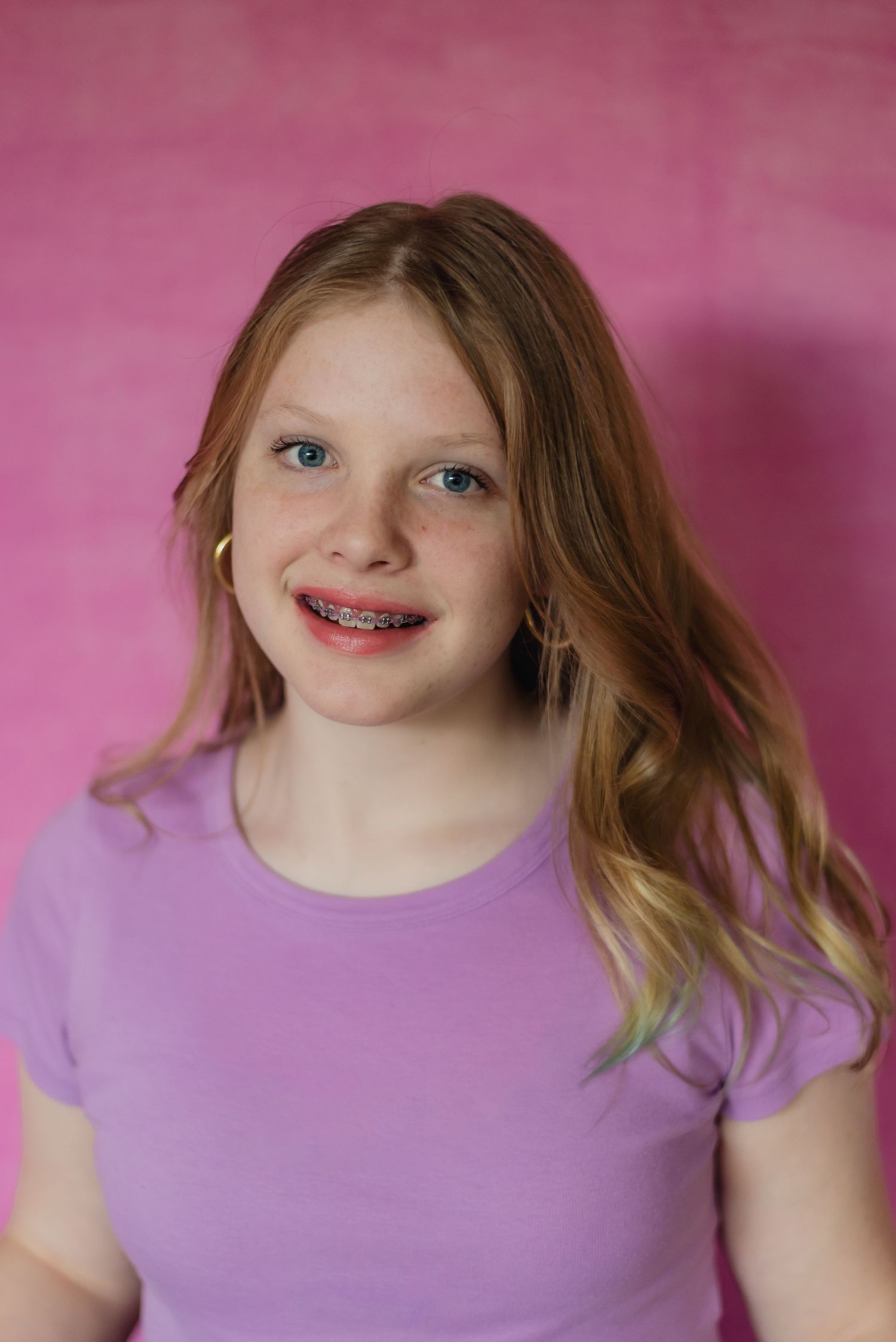 Young teen girl wearing a purple t-shirt smiles at camera against pink background, showing braces. Headshot photography in Snohomish County.