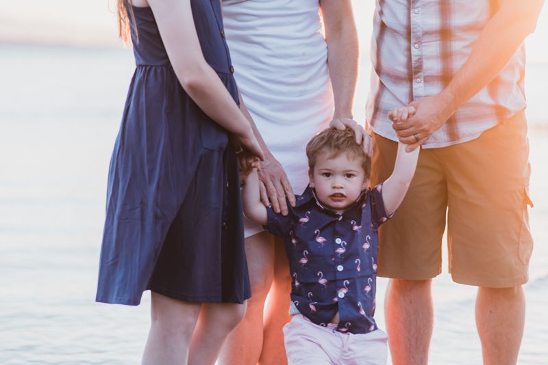 A toddler in a navy polka dot dress walks along a beach at sunset while holding an adult's hand.