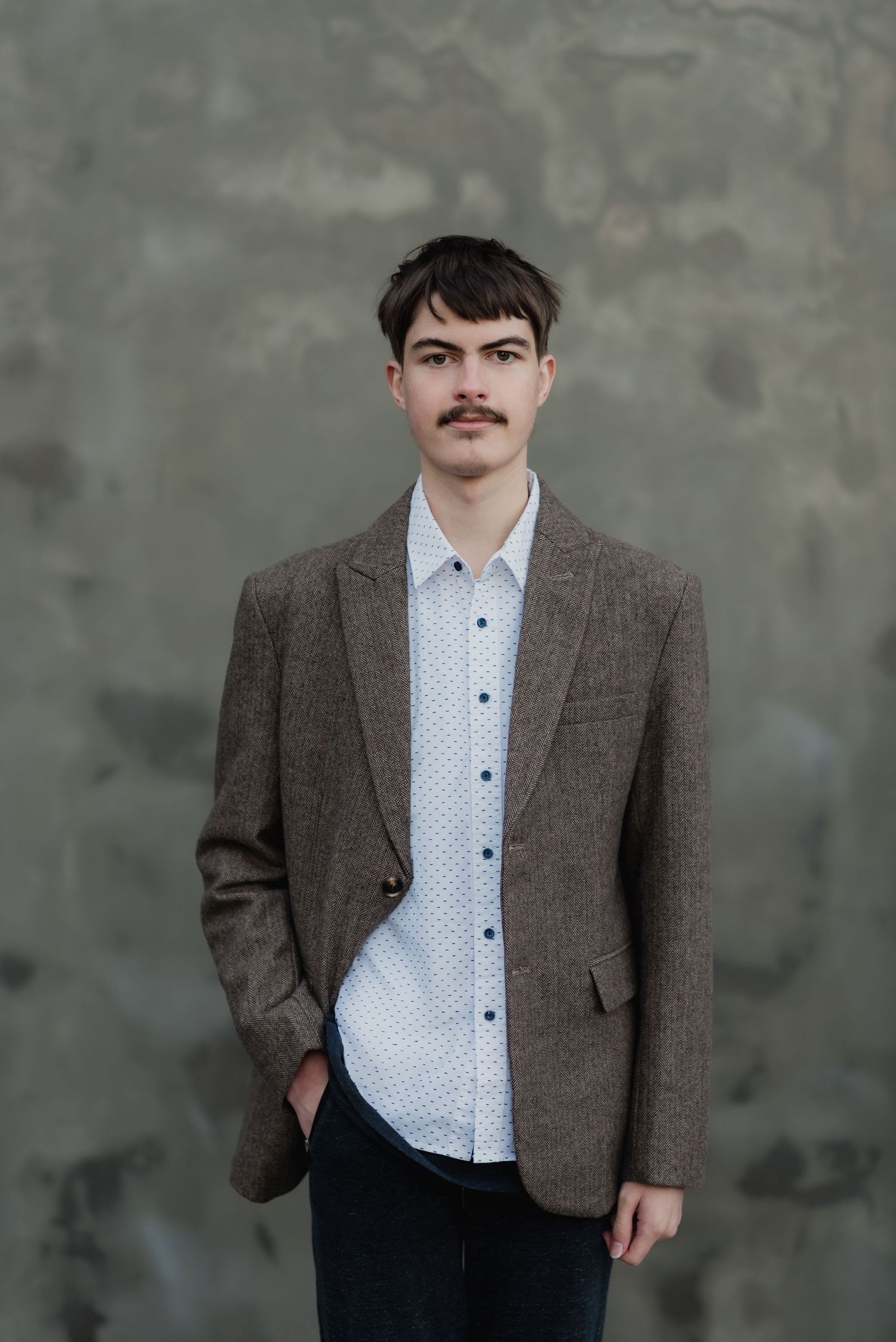 Young man with mustache wearing brown tweed blazer and white dotted shirt standing against gray wall.