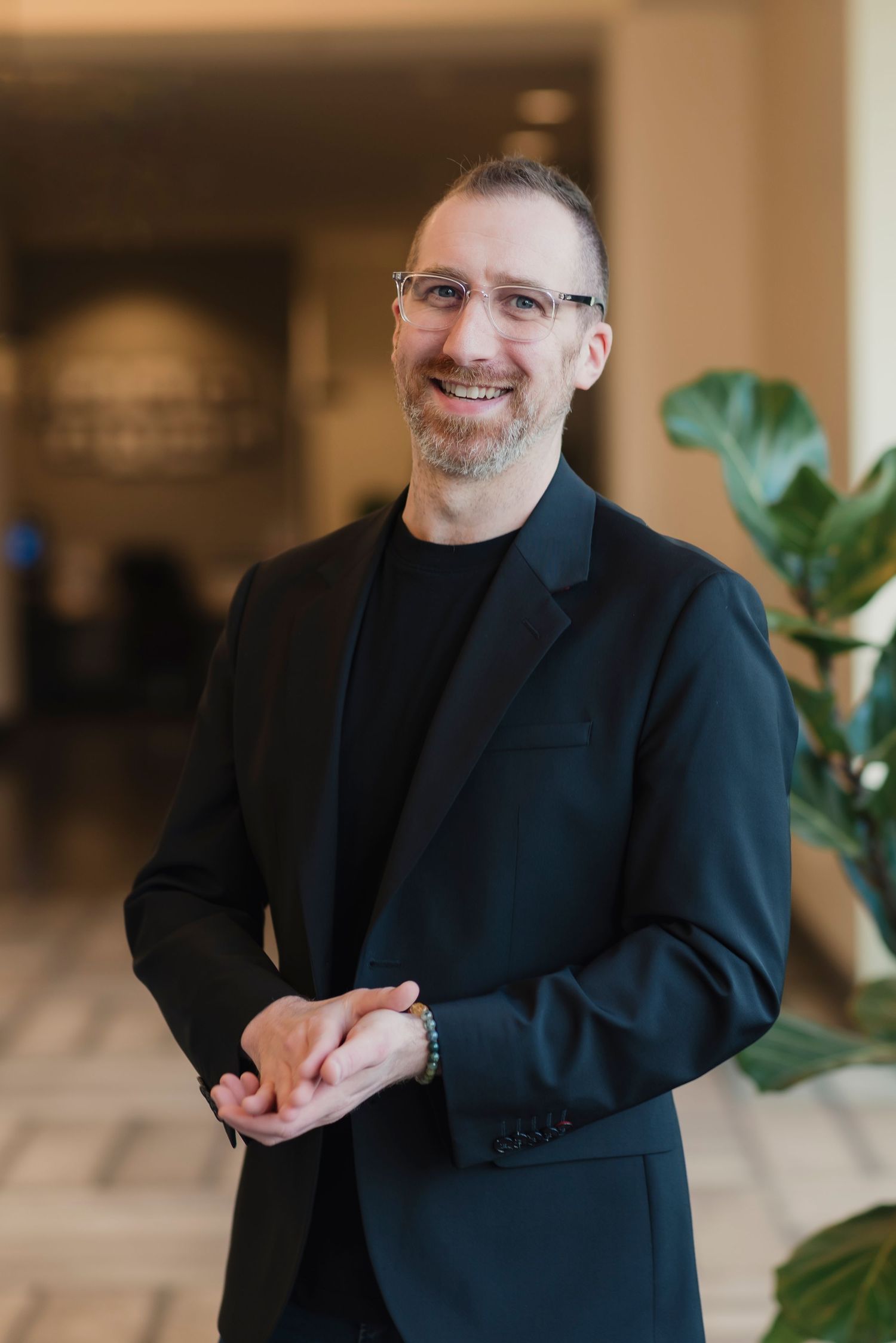 Professional man in black blazer smiling confidently in modern lobby setting with glasses and neatly groomed beard.