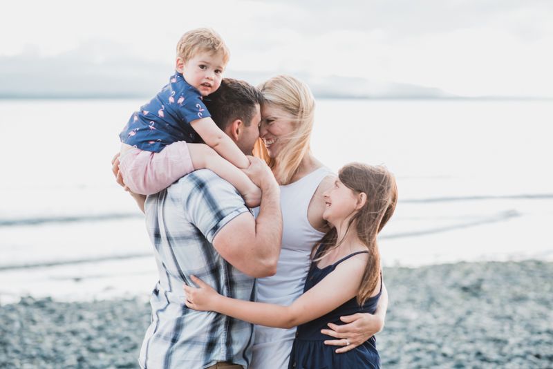 A loving family embraces together on a peaceful beach with mountains and water in the background.