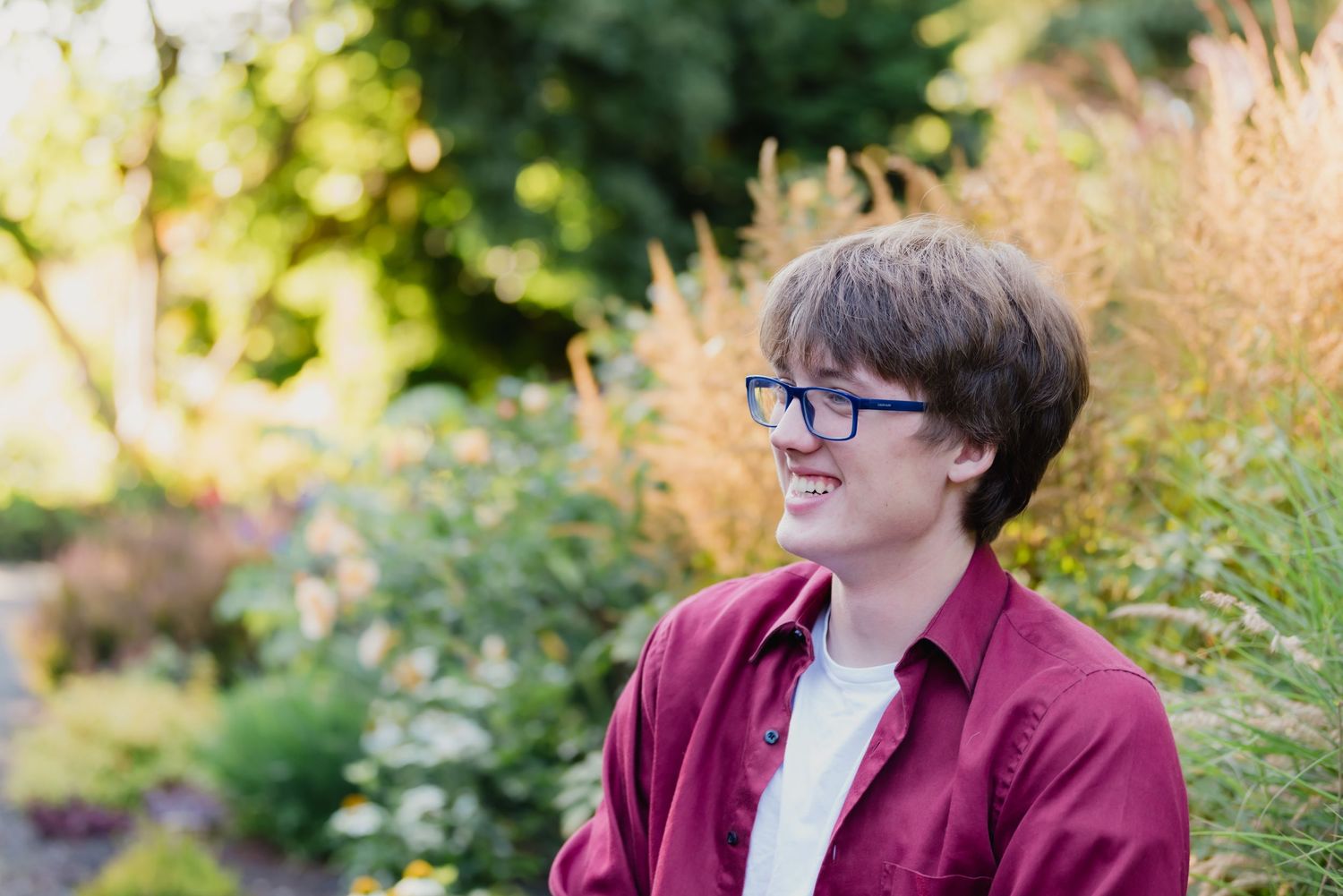 A person wearing glasses and a burgundy cardigan sits outdoors among lush green foliage.