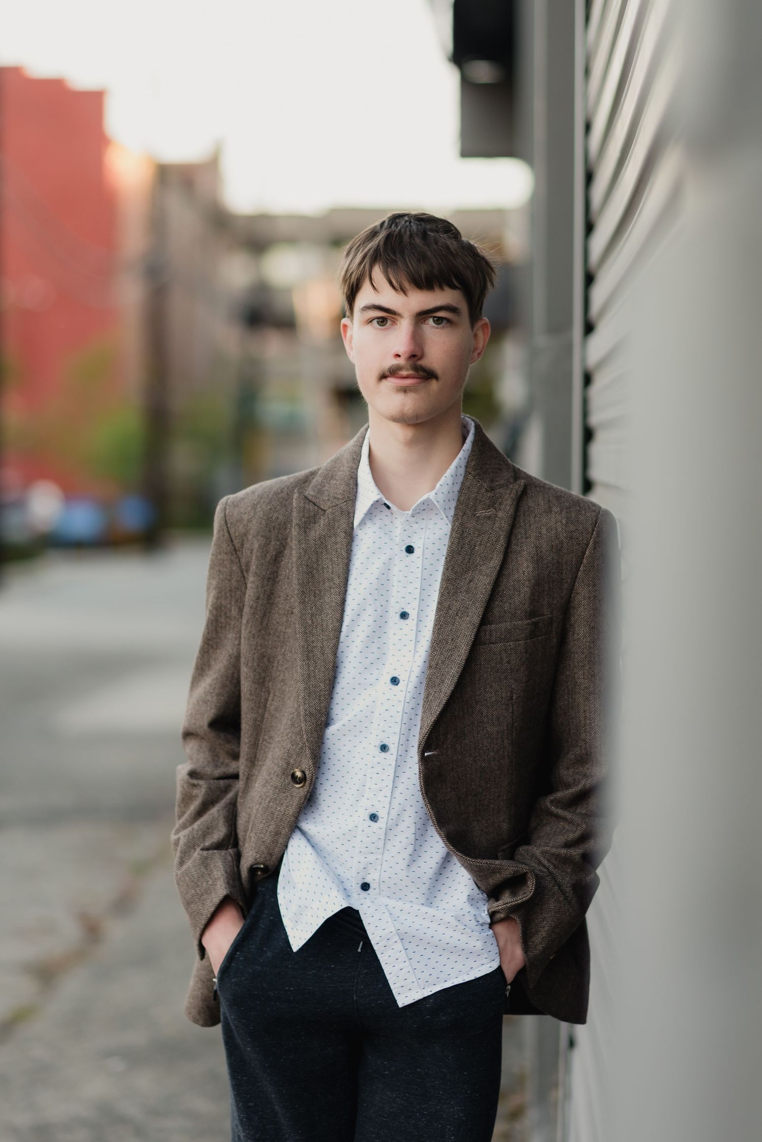 Young man in brown tweed blazer and polka dot shirt poses against column in urban setting.