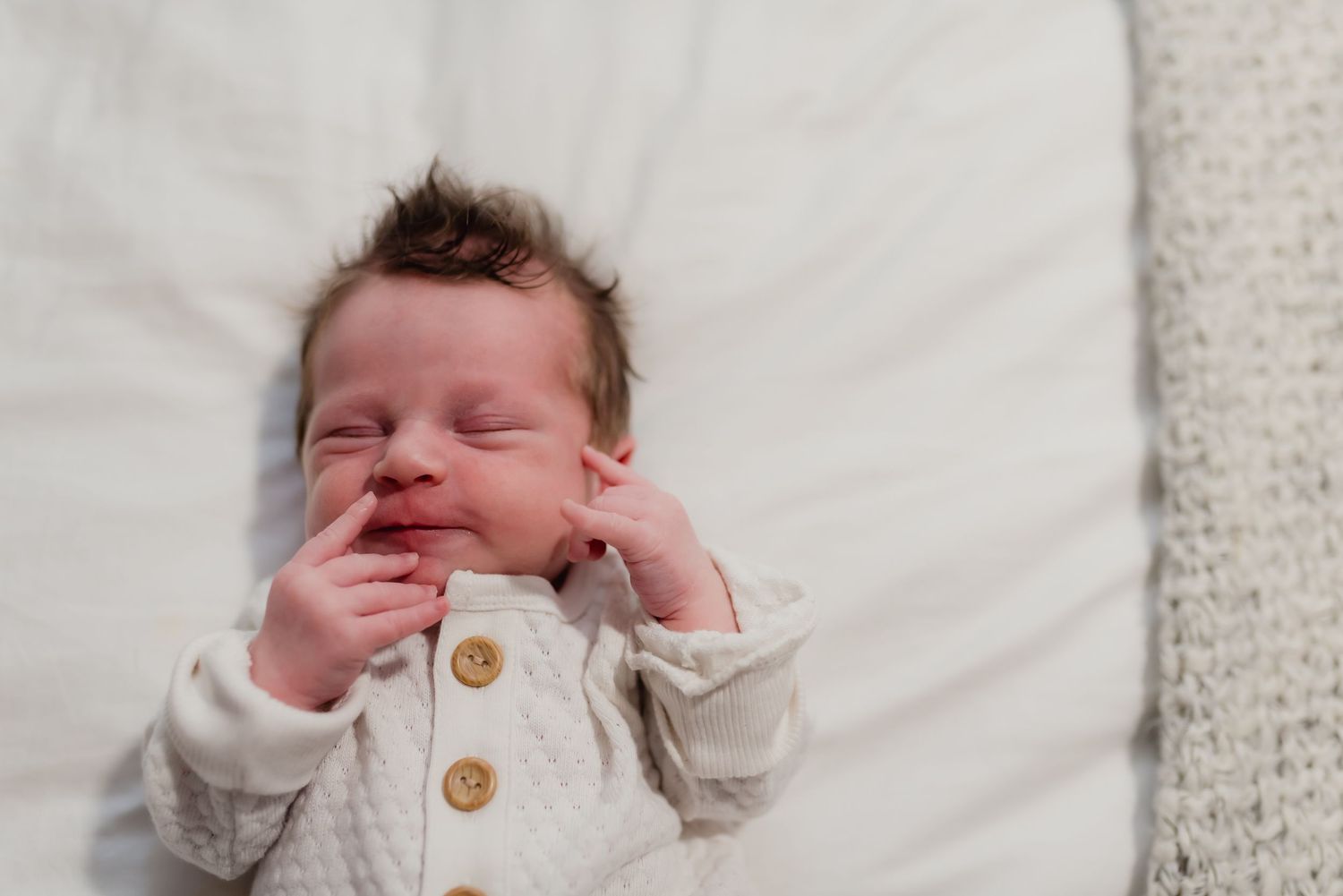 A sleeping baby lies on white bedding wearing a cozy oatmeal-colored sweater with wooden buttons.