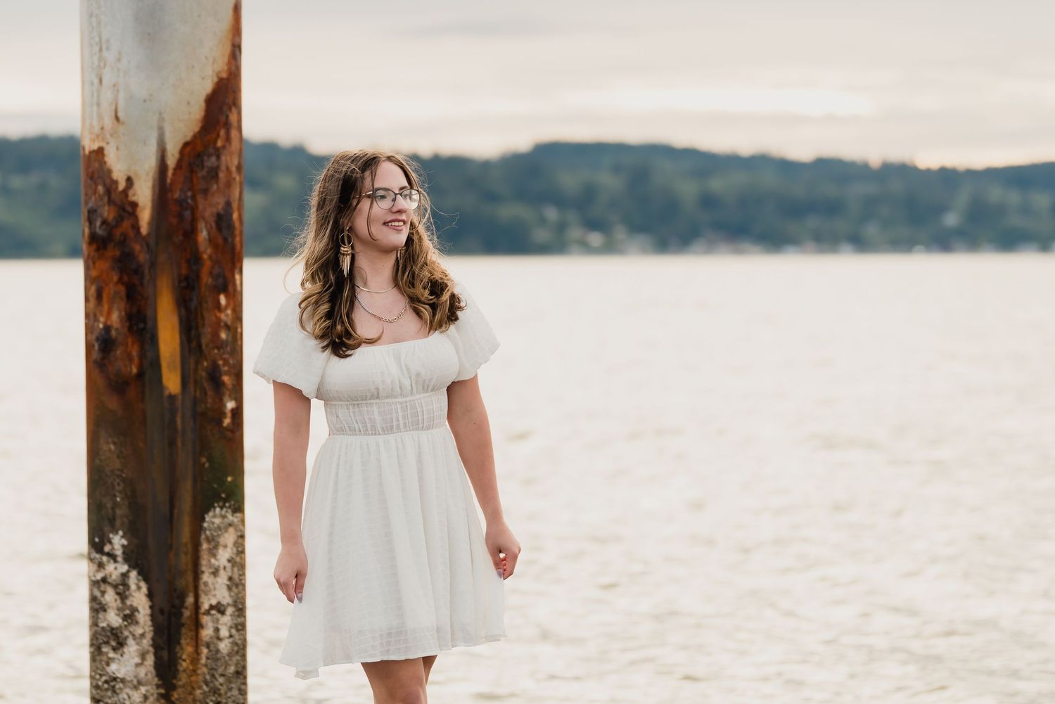 A person in a white dress stands near rustic wooden posts along a serene waterfront at sunset.