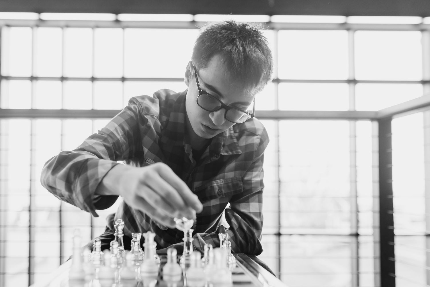 Black and white photo of a young man wearing glasses playing chess near a large window.
