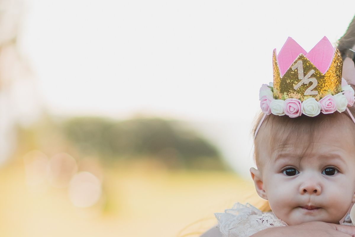 Baby wearing a floral crown headband and white dress smiles in outdoor portrait.