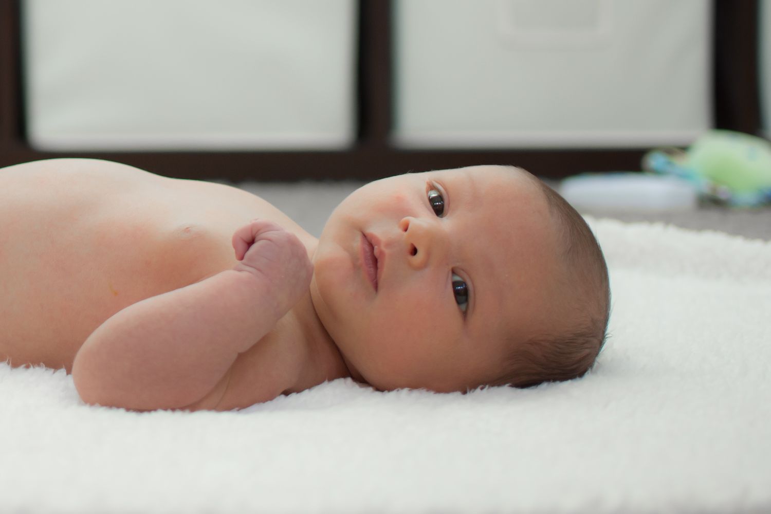 A newborn baby lies peacefully on white bedding while looking up with wide eyes.