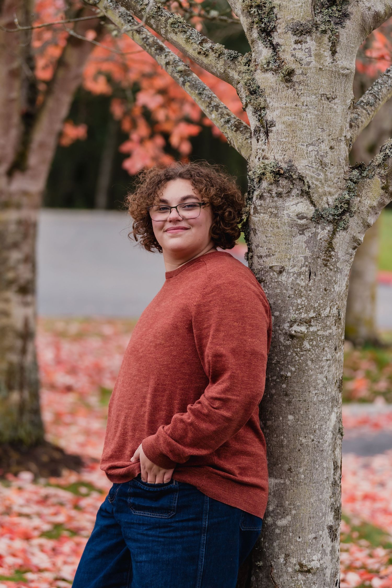 Person in a rust-colored sweater and jeans poses by a tree trunk with autumn leaves in the background.