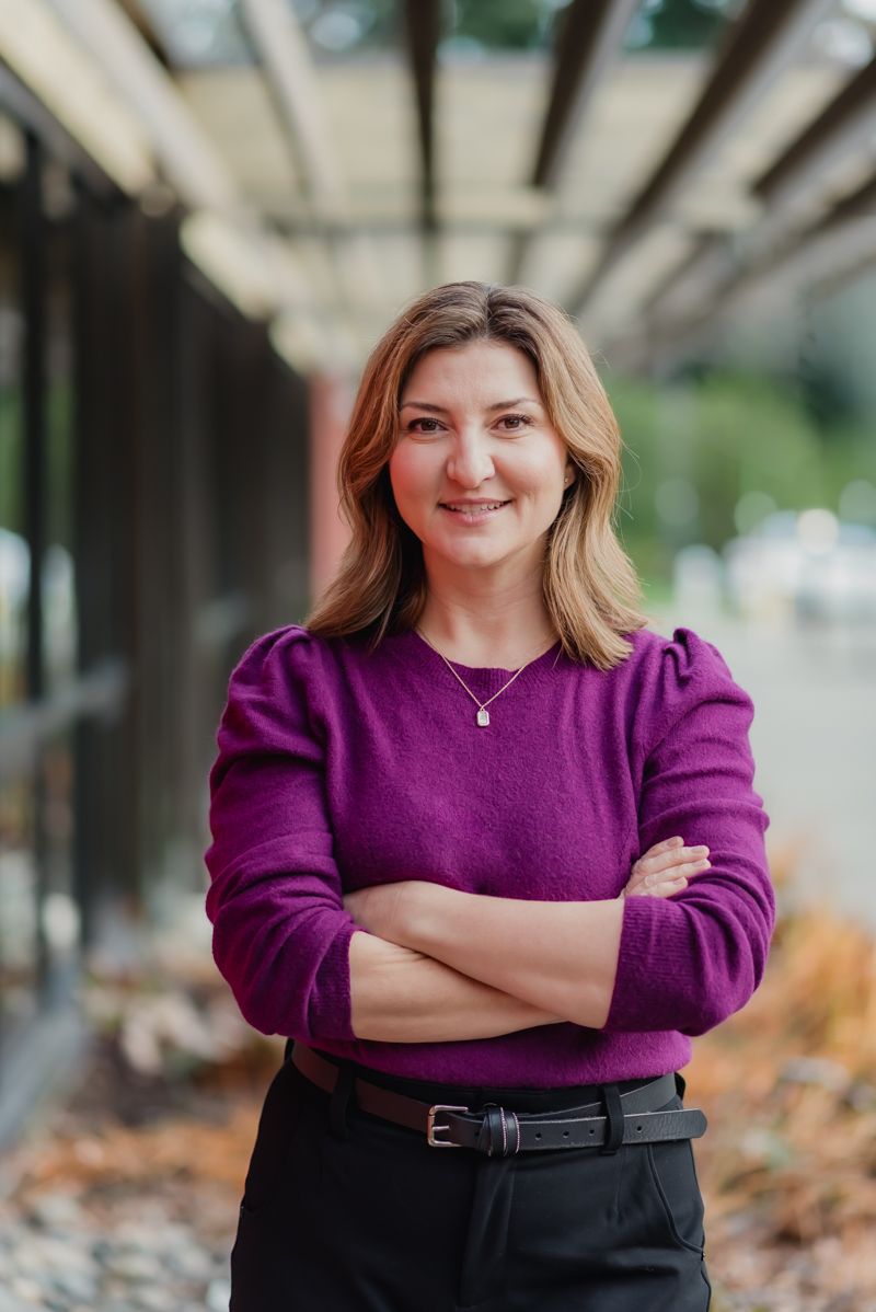 Professional woman in purple sweater smiling with arms crossed outdoors near modern building.