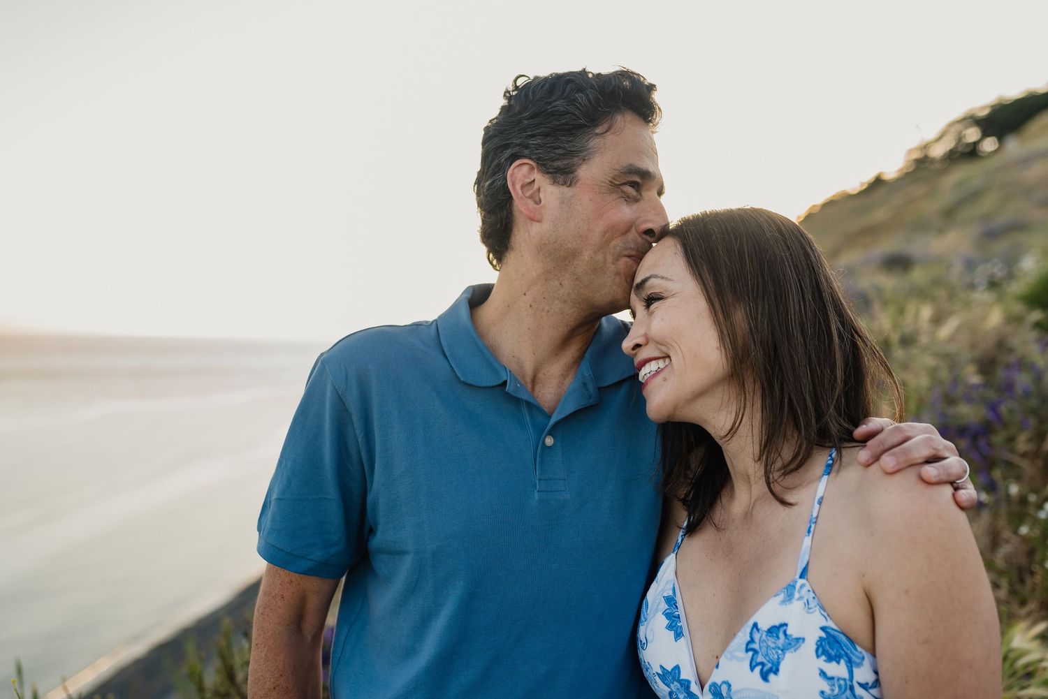 A couple in blue attire shares romantic moments during a coastal photo session at sunset.