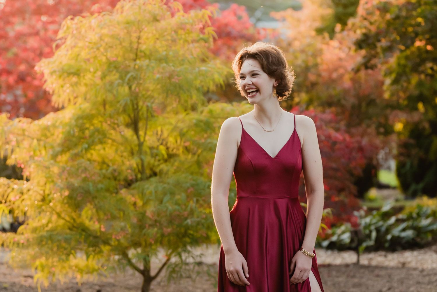 High School Senior girl in a long burgundy prom dress smiles while standing in front of vibrant yellow and red autumn foliage in Everett, WA