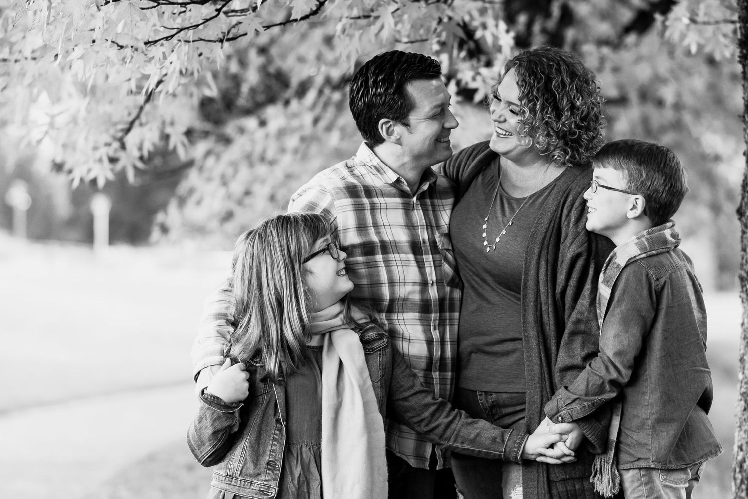 family of four holding hands and looking at each other while standing under a tree