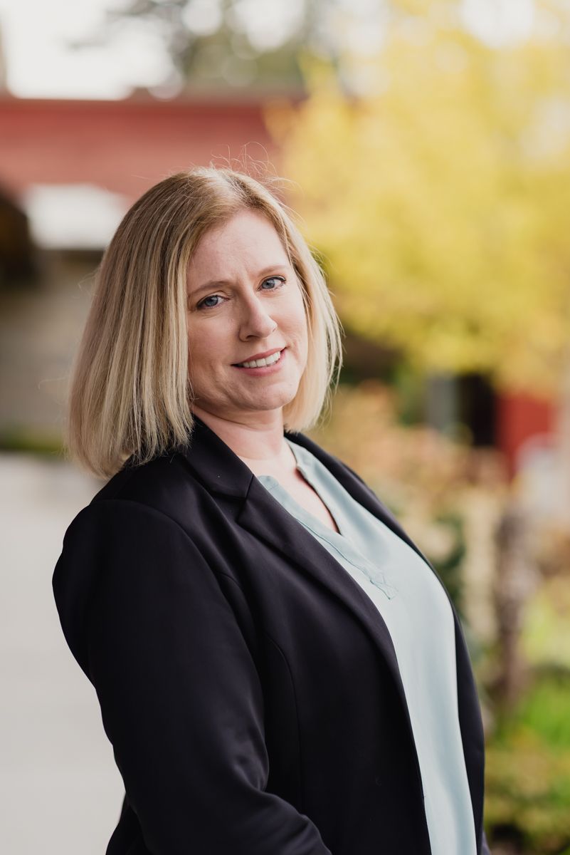 Professional woman in black blazer and light blue blouse smiling outdoors with autumn foliage background.