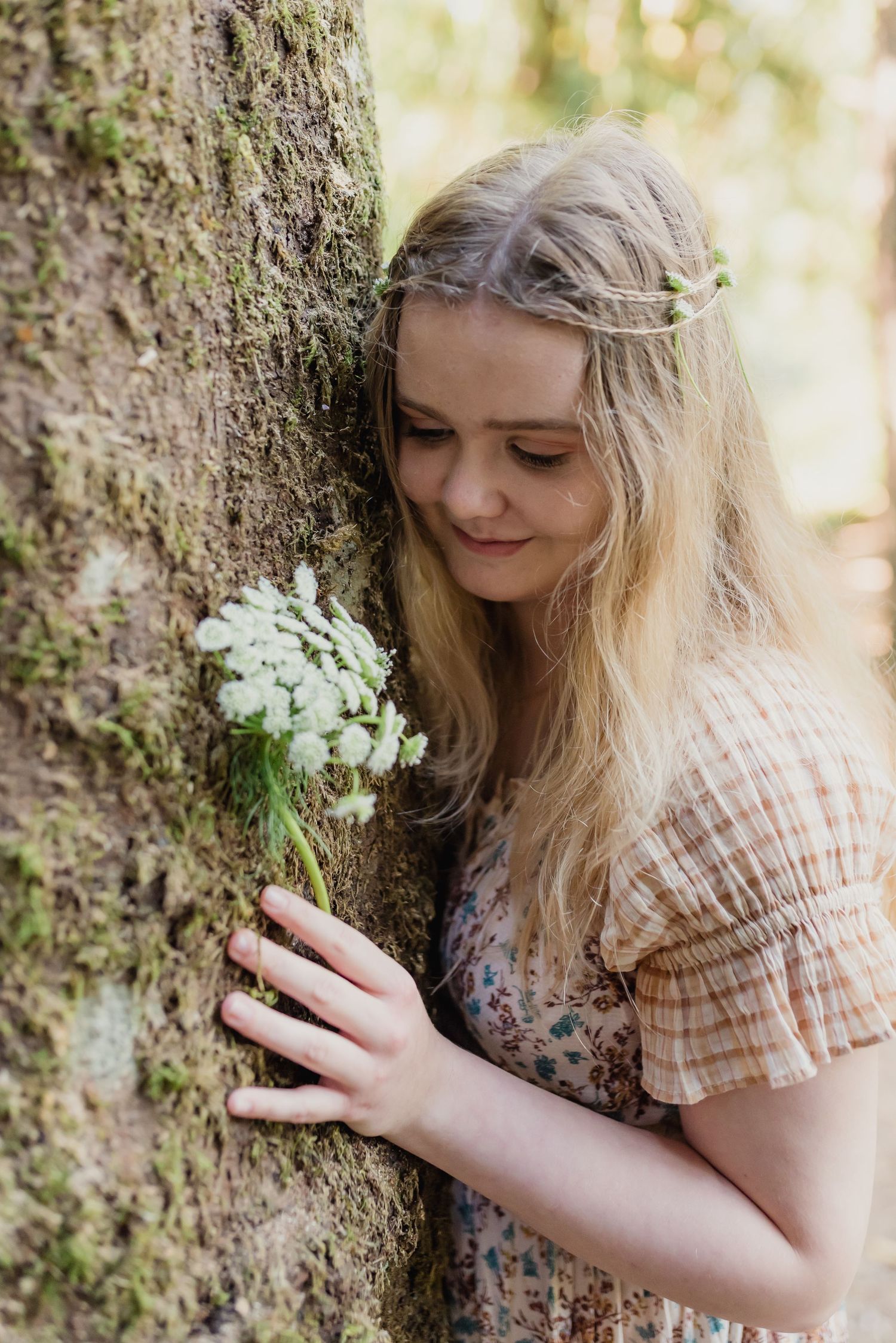 A dreamy forest photoshoot series shows a person in floral dress examining moss and lichen on a tree trunk.