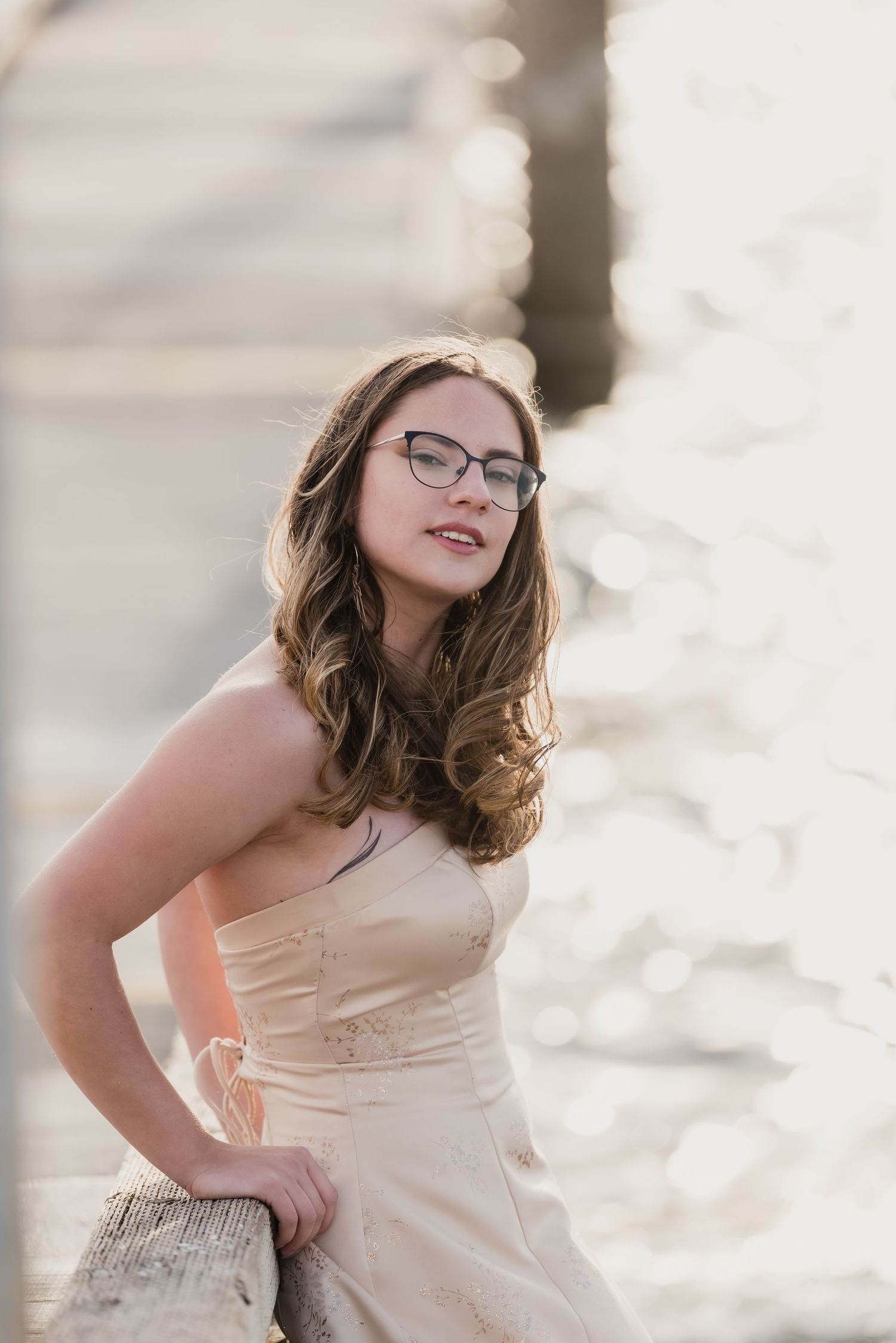 A high school senior girl in glasses and a cream-colored dress leans on the dock during golden hour at Mukilteo Lighthouse Park.