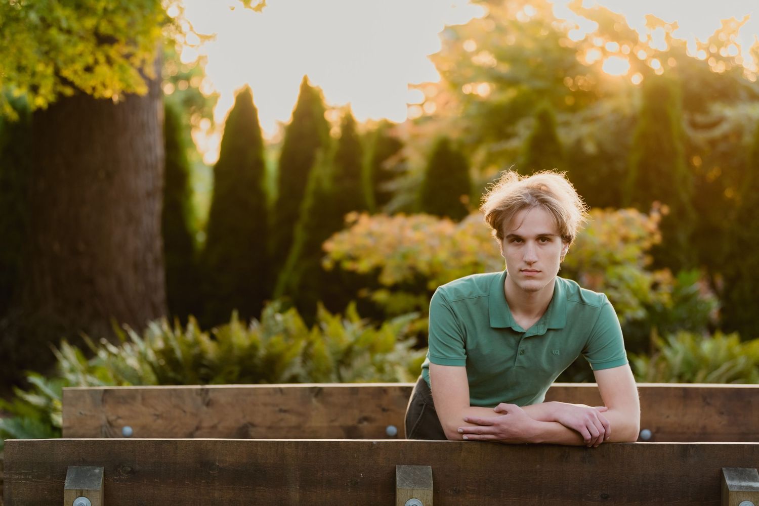 Person in green polo shirt sitting at wooden bench in garden with lush greenery and sunset lighting in background.