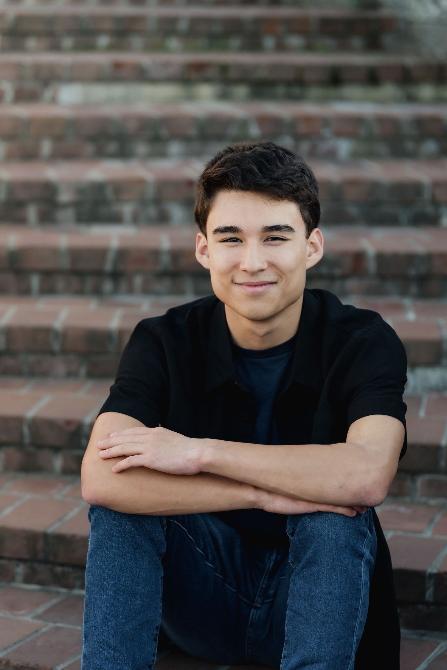 Mukilteo Kamiak High School senior young man in black shirt and jeans sitting on brick steps with arms crossed, smiling confidently.