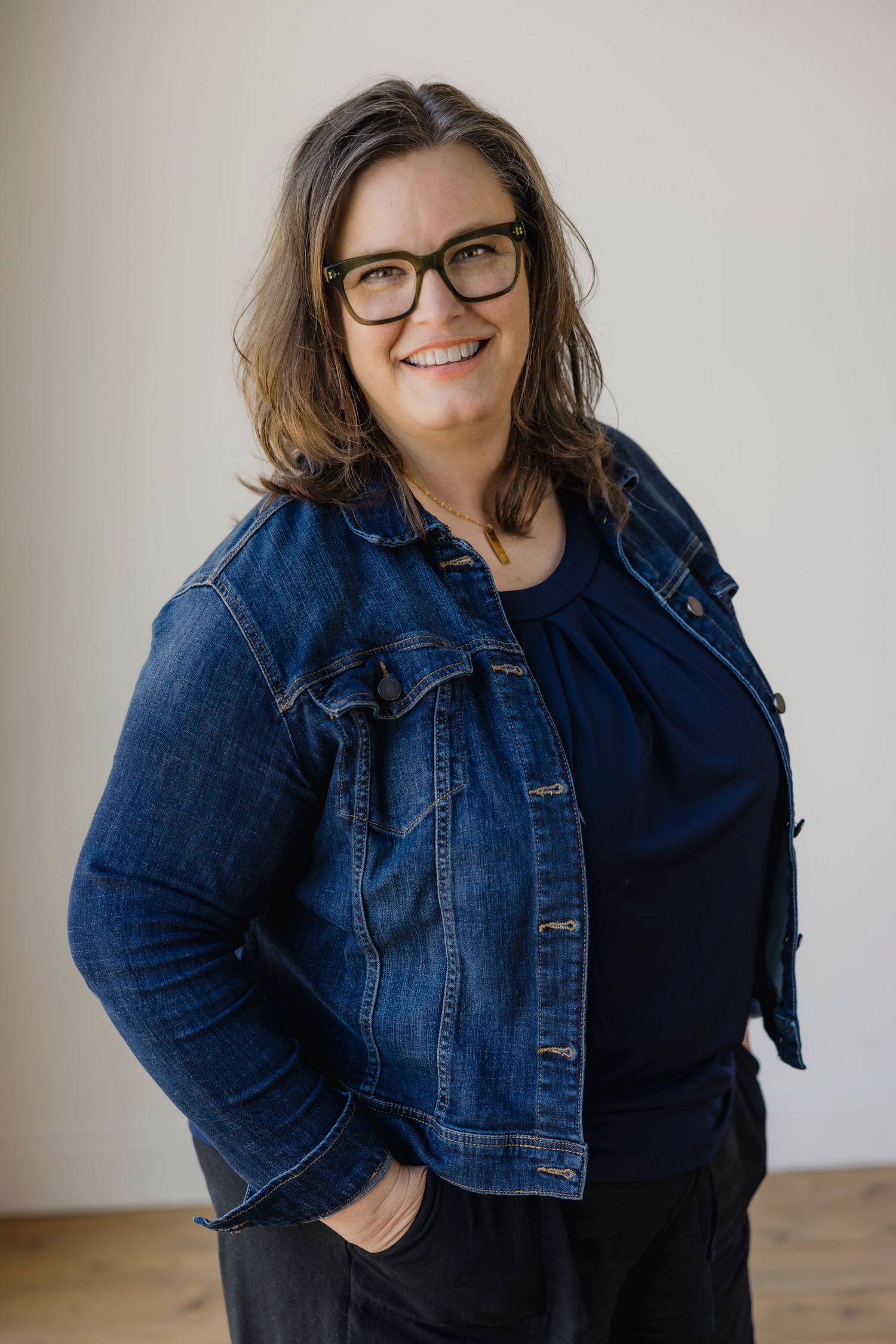 Smiling woman with glasses wearing a denim jacket and dark top, posing with hands in pockets against a neutral background.