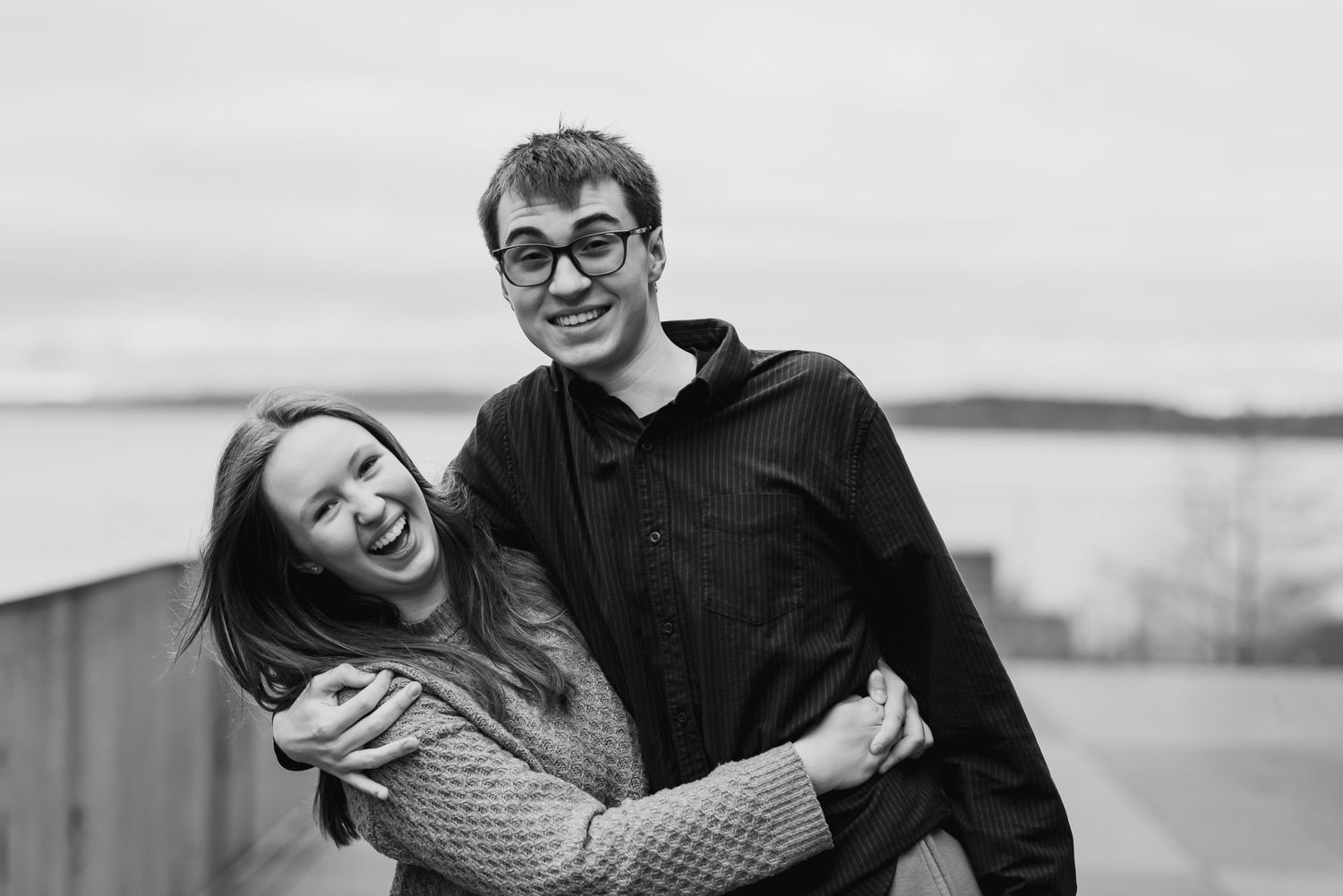 Black and white photo of a smiling couple embracing outdoors near a waterfront on a cloudy day.