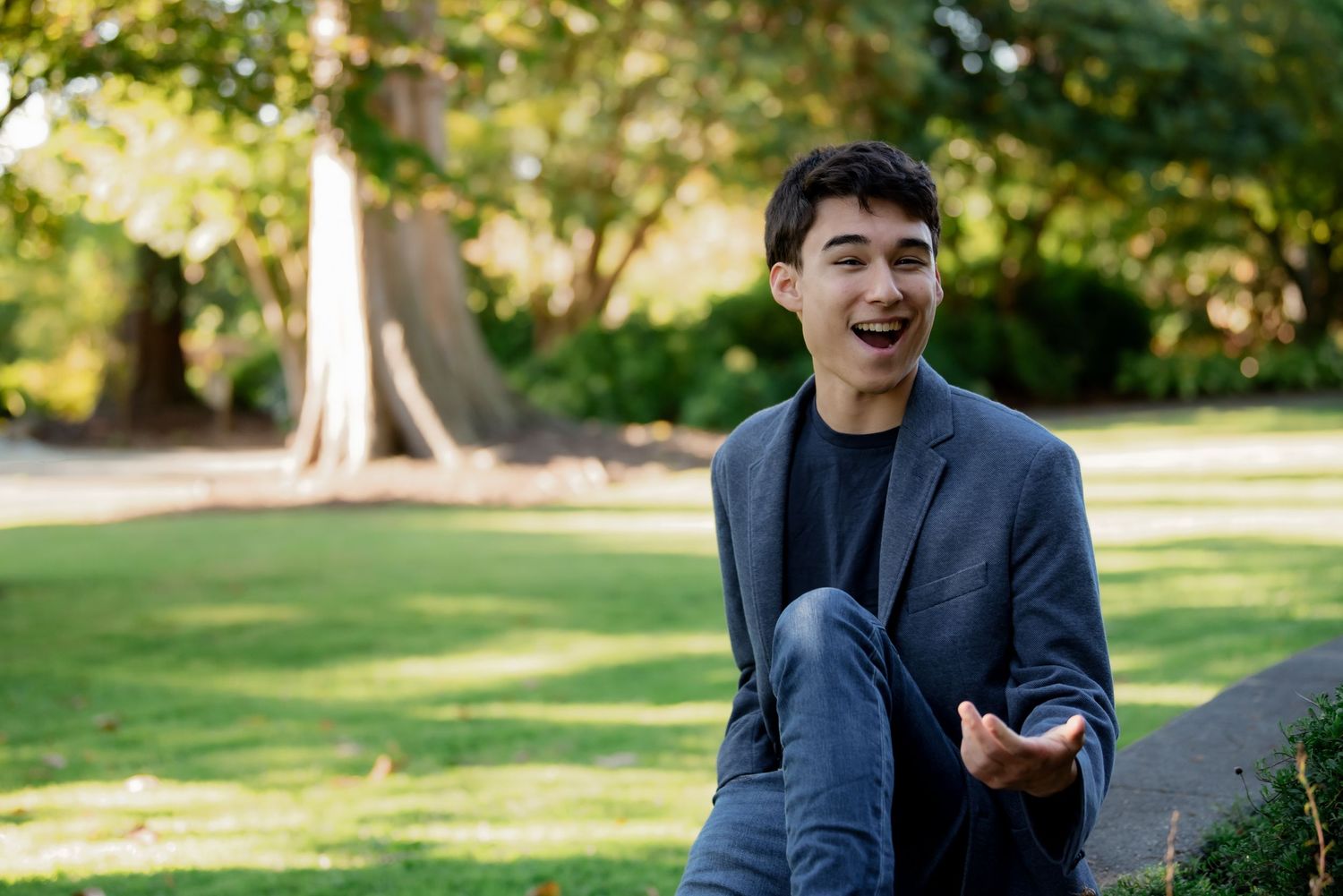 A person wearing a navy blazer smiles while holding a phone outdoors in a sunny park setting.