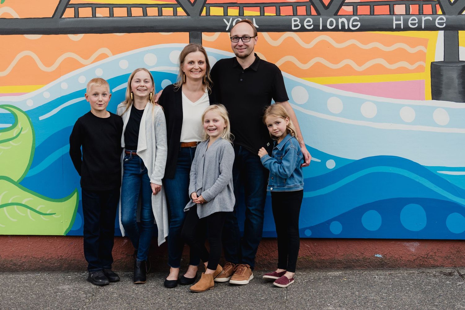Family of 6 with three girls and one boy stands in front of colorful mural with wavy lines and patterns that says You Belong Here.