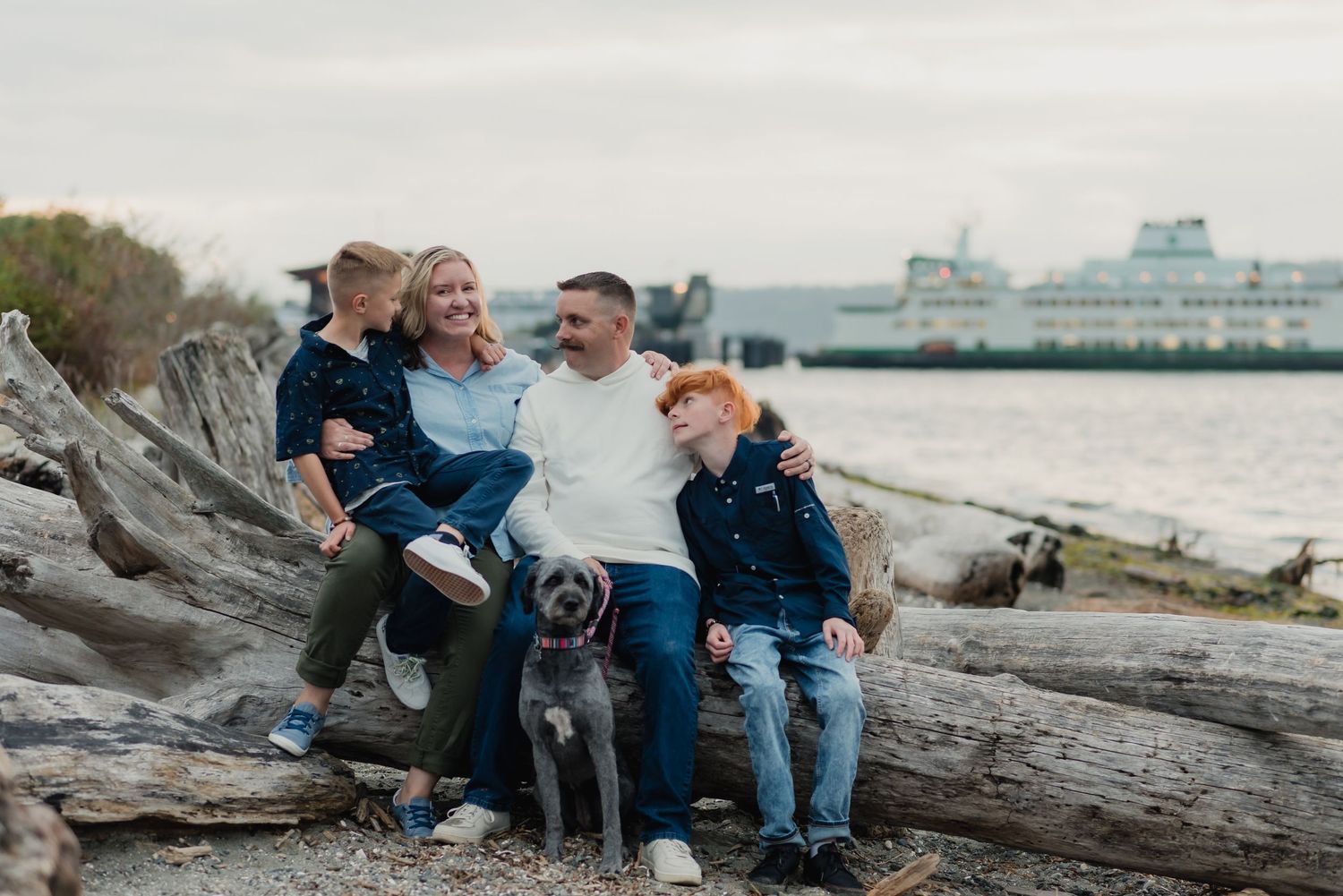 A group sits together with their dog on driftwood at the beach with a ferry visible in the background.