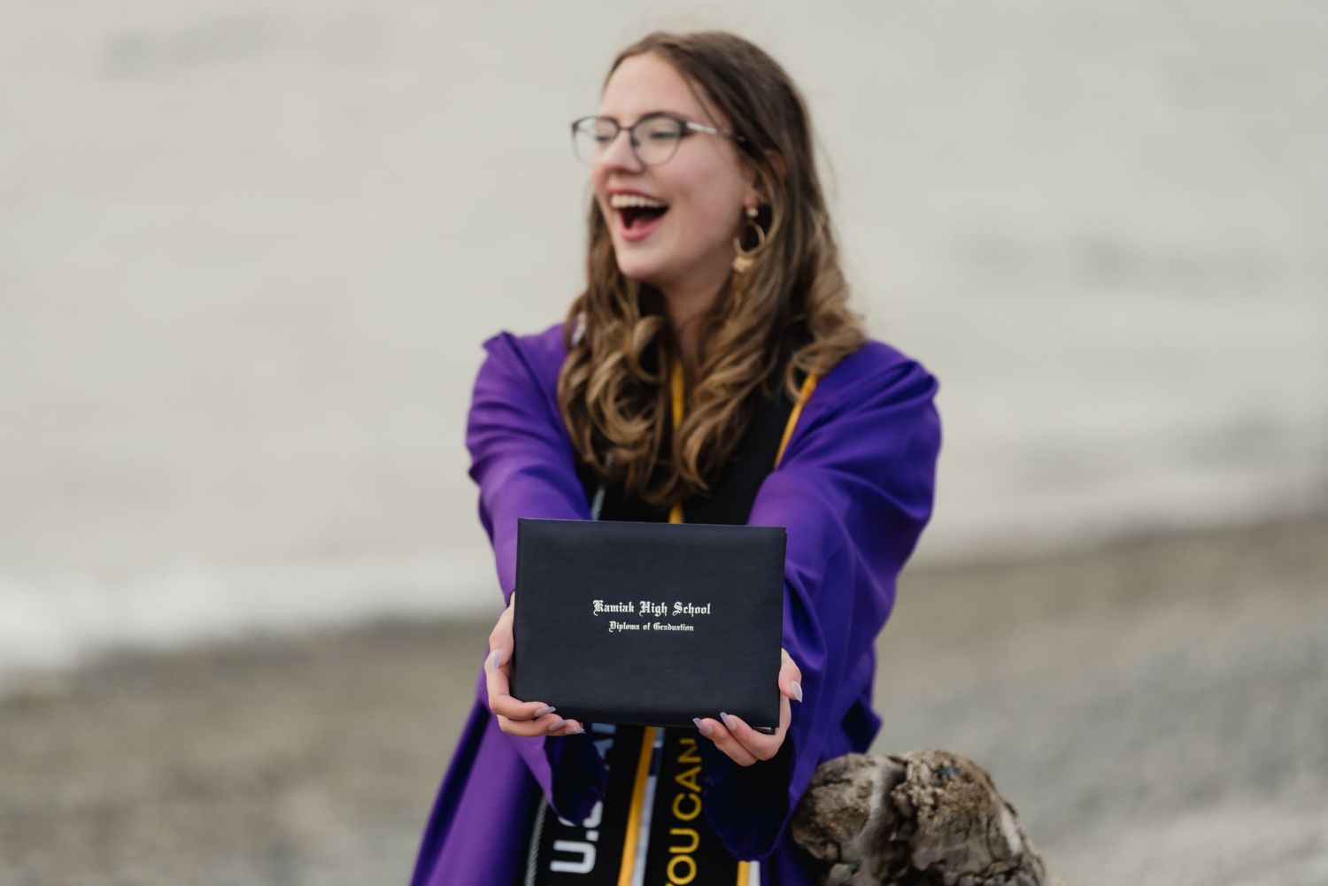 A joyful graduate in a purple gown holds their diploma while laughing outdoors on a cloudy day.