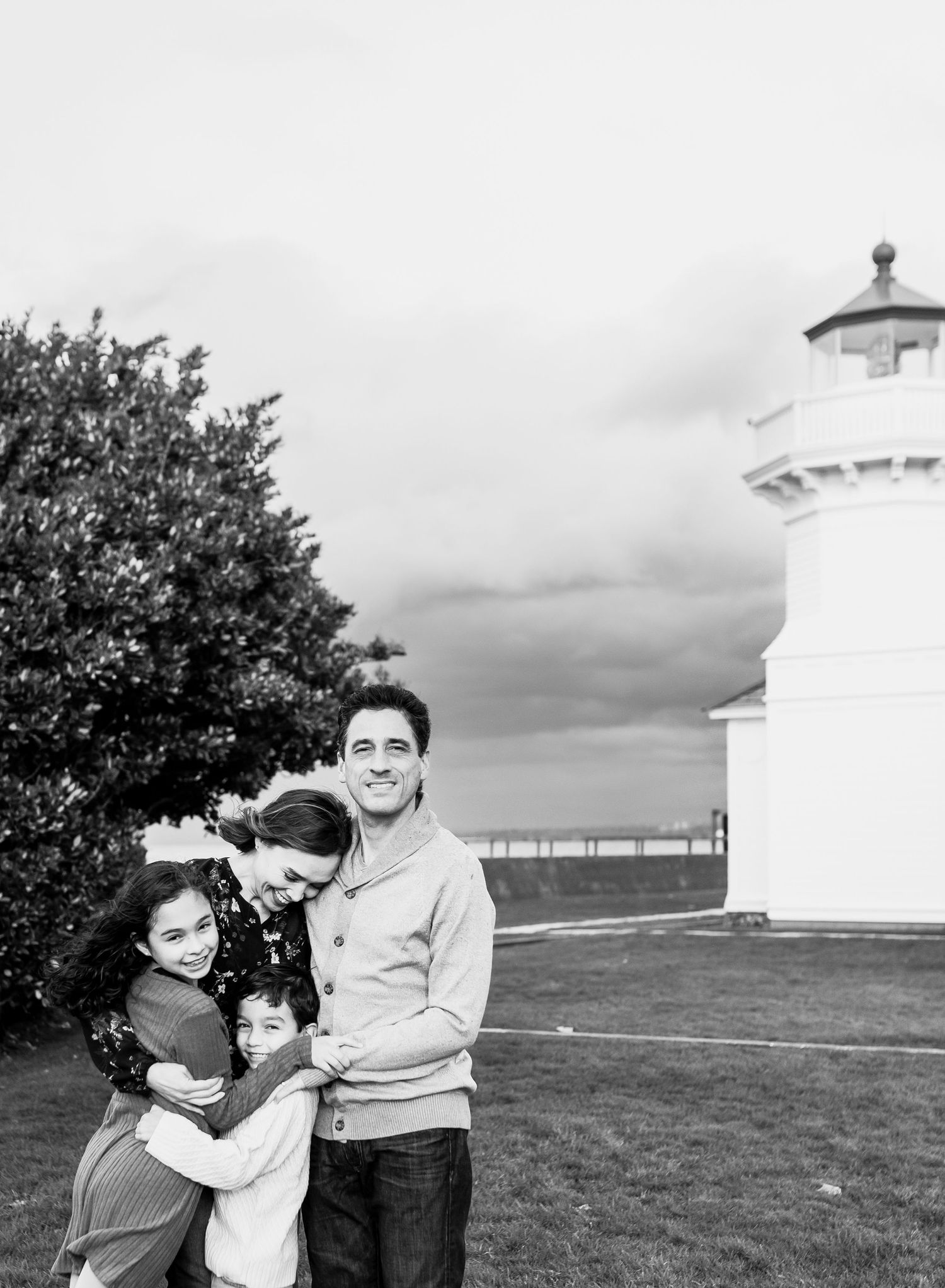 Black and white family portrait near a lighthouse, parents embracing three children on a grassy lawn.