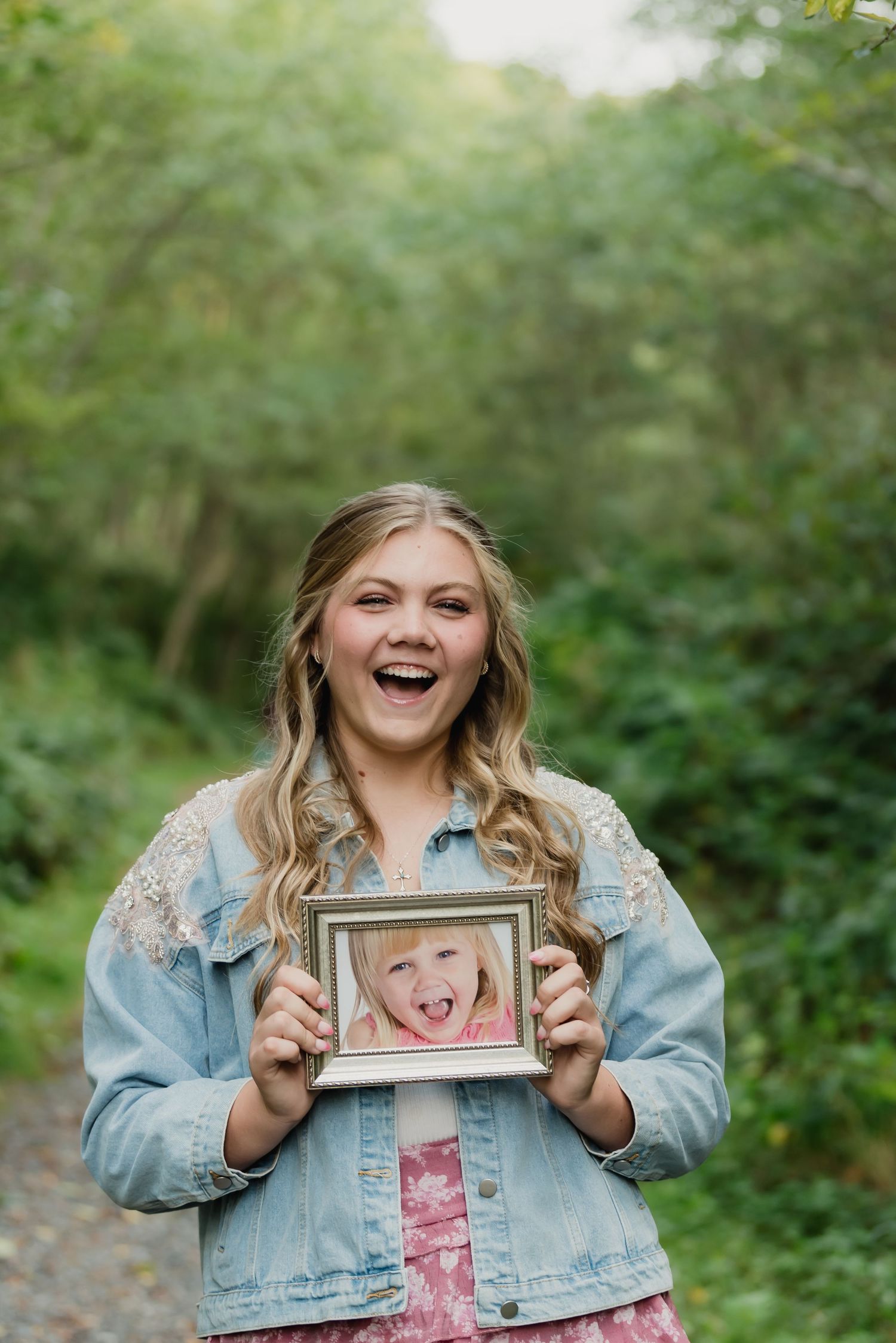 A person wearing a denim jacket holds up a photo frame while laughing joyfully in a green outdoor setting.