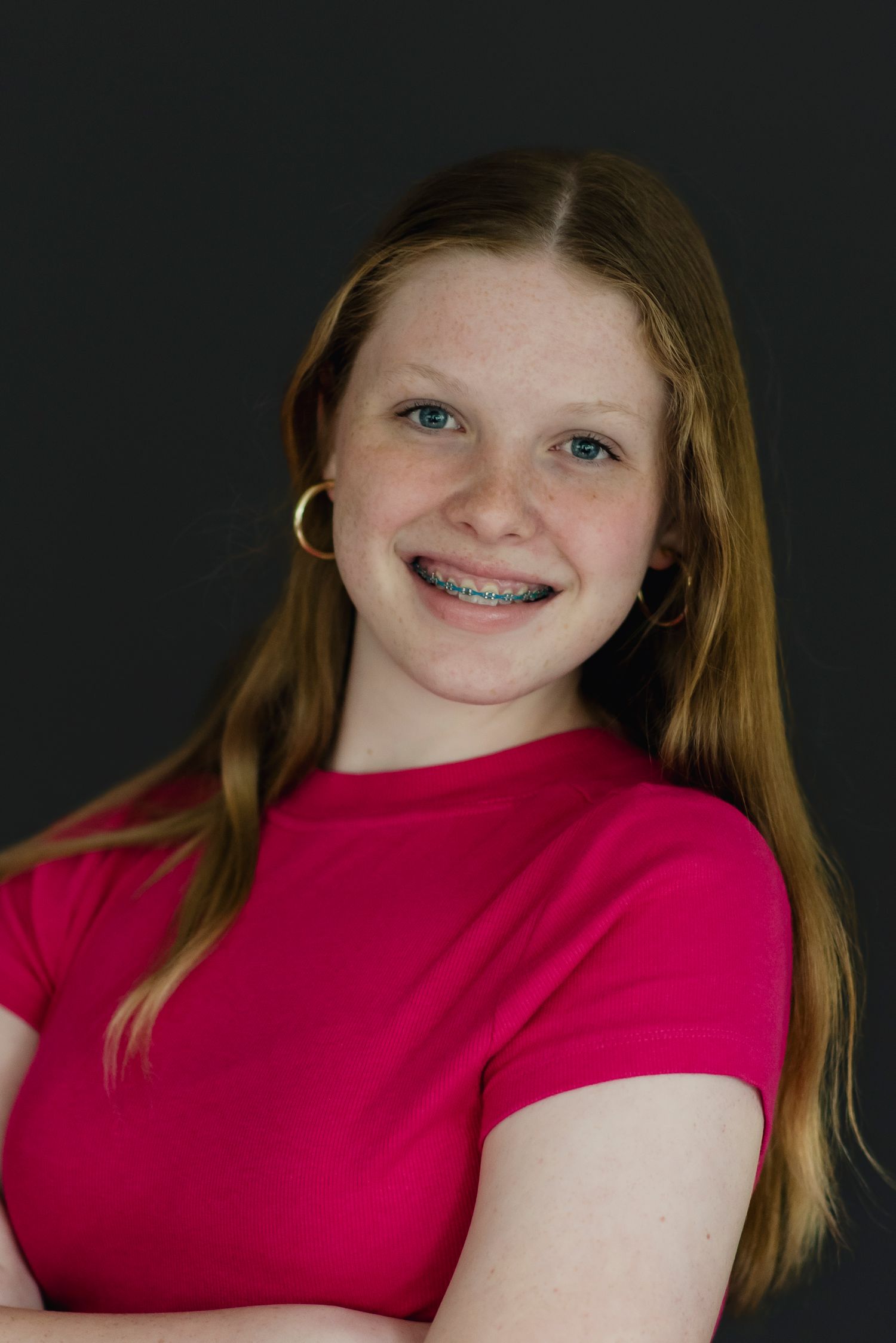 Smiling teenage girl with braces and long red hair wearing a pink top and gold hoop earrings against dark background.