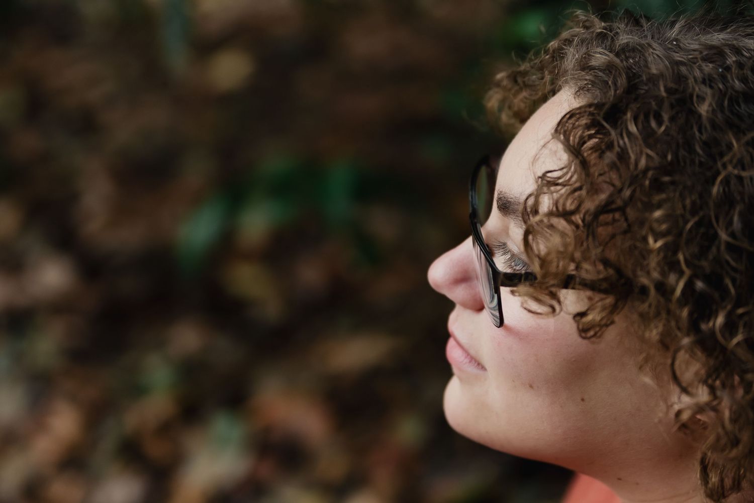 A person with curly hair and glasses gazes upward in a natural outdoor setting with blurred foliage in background.