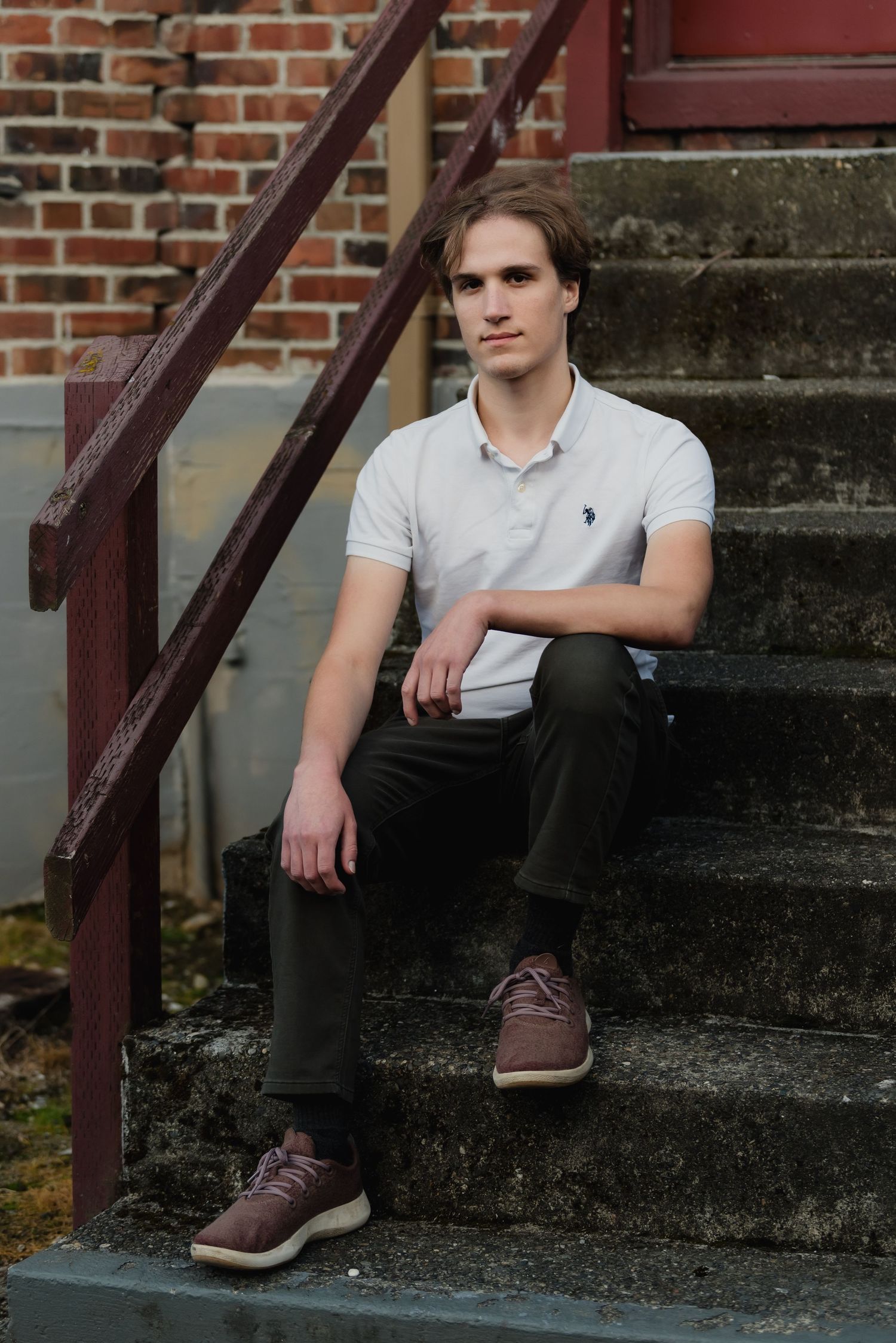 Someone in a casual gray polo shirt and dark pants sits on concrete steps near a brick wall with a metal railing.