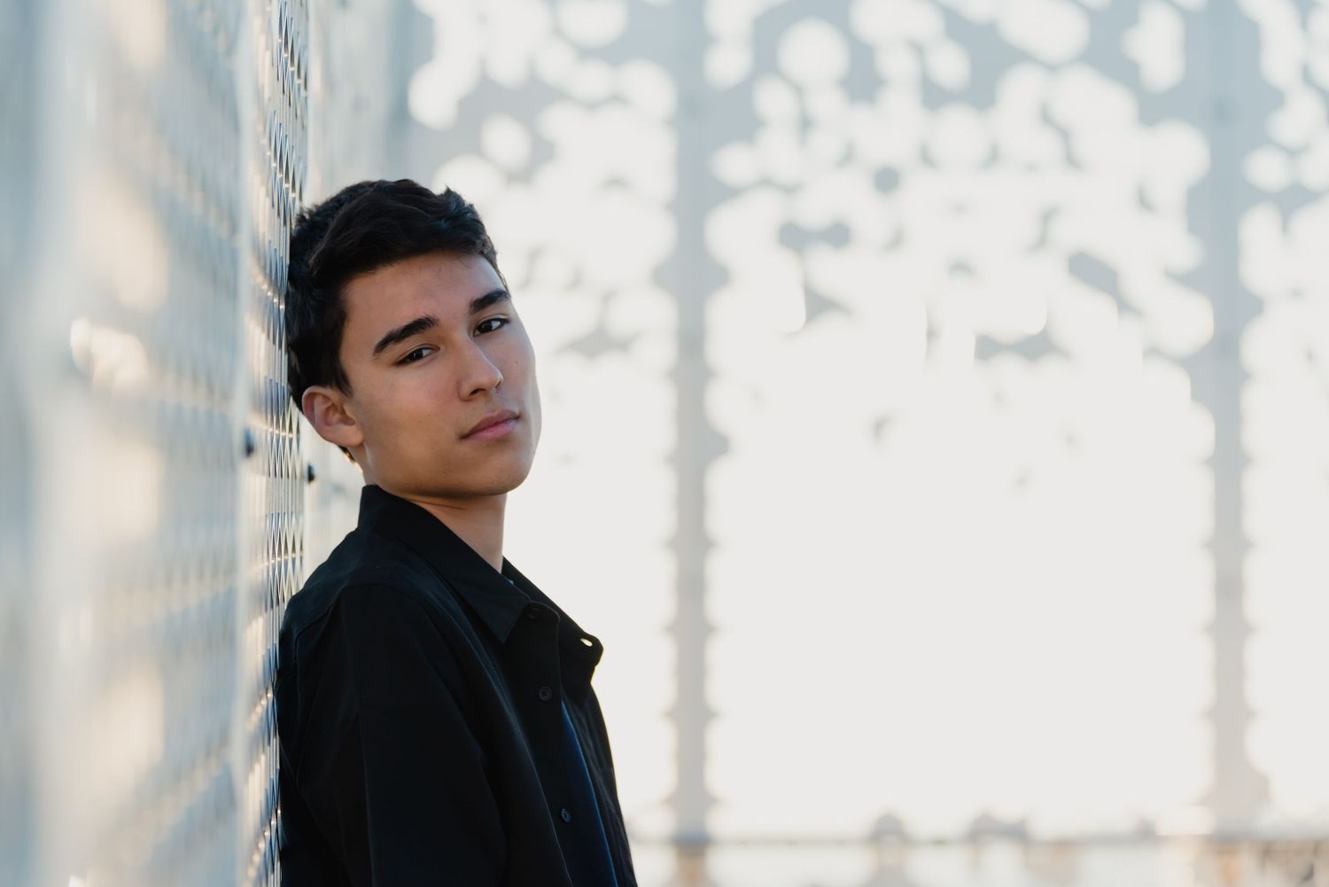 Person in black jacket poses against light-filled wall with dramatic shadows and soft lighting effect.