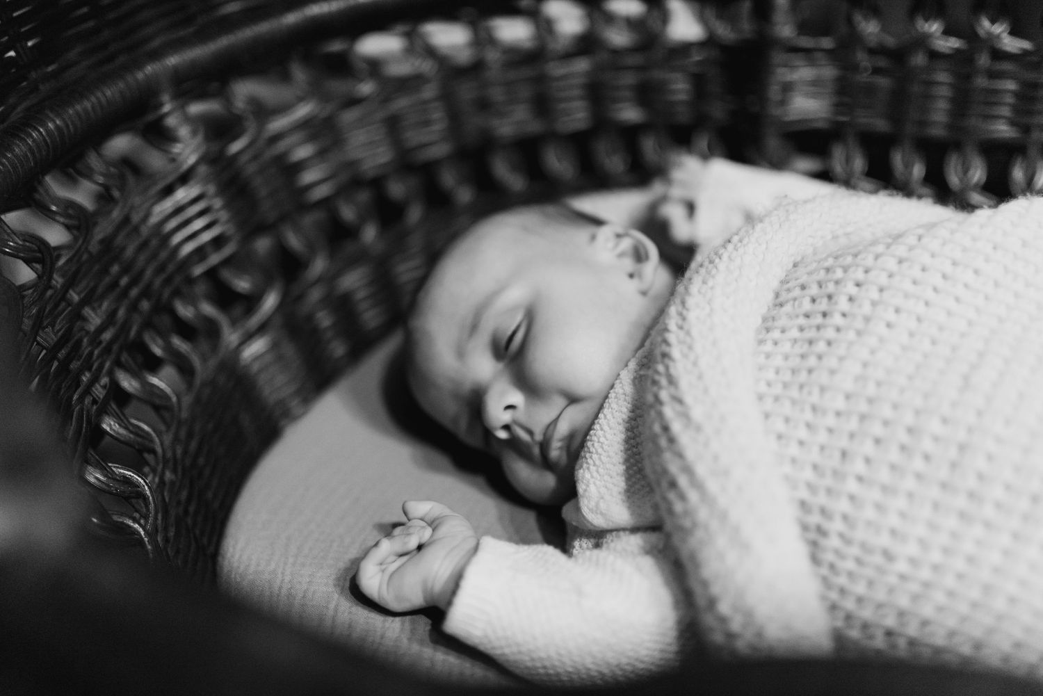 A sleeping baby in a white knit sweater rests peacefully in a woven basket, captured in black and white photography.