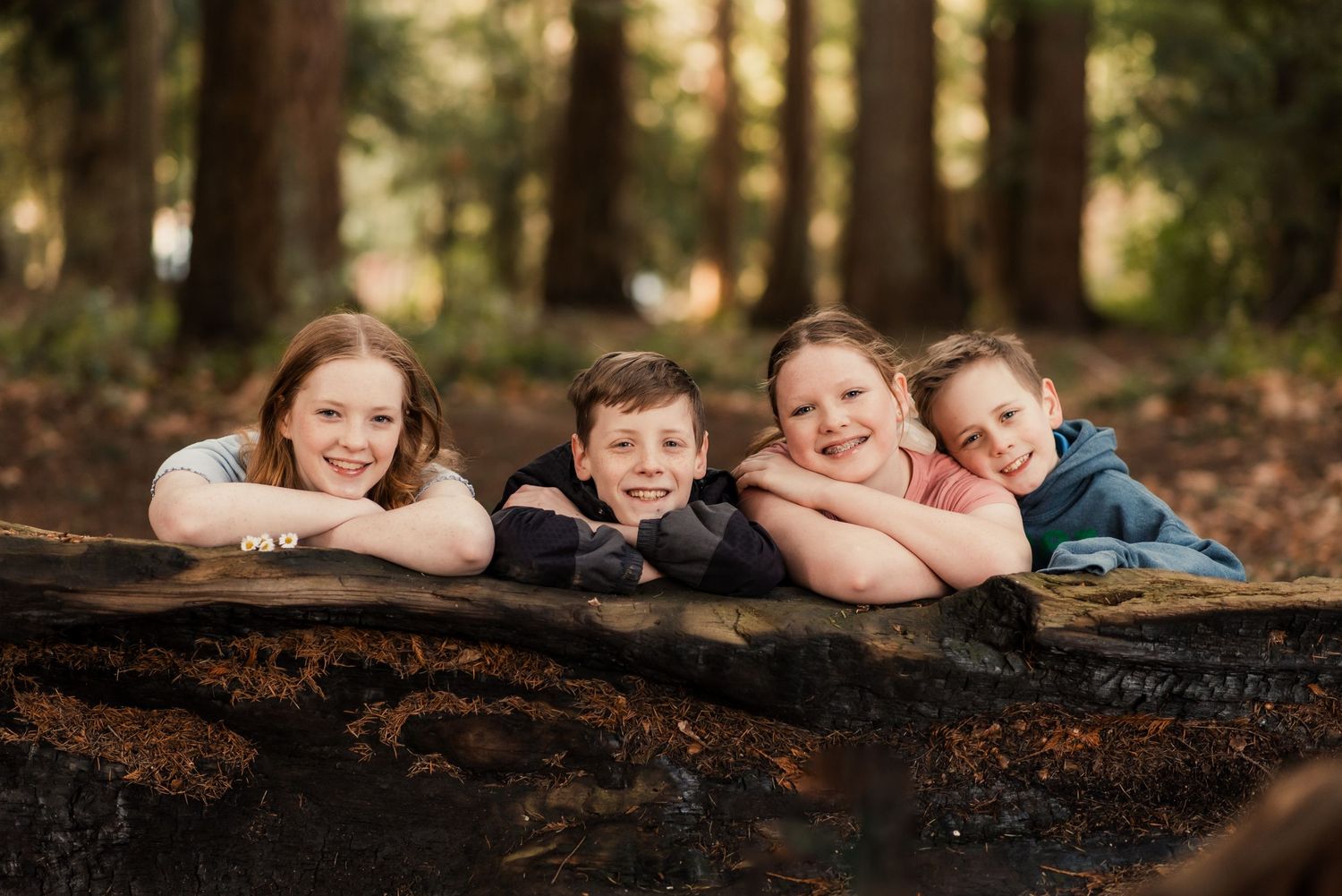 A group of children laying on a fallen log in a sunlit forest setting, smiling at the camera.