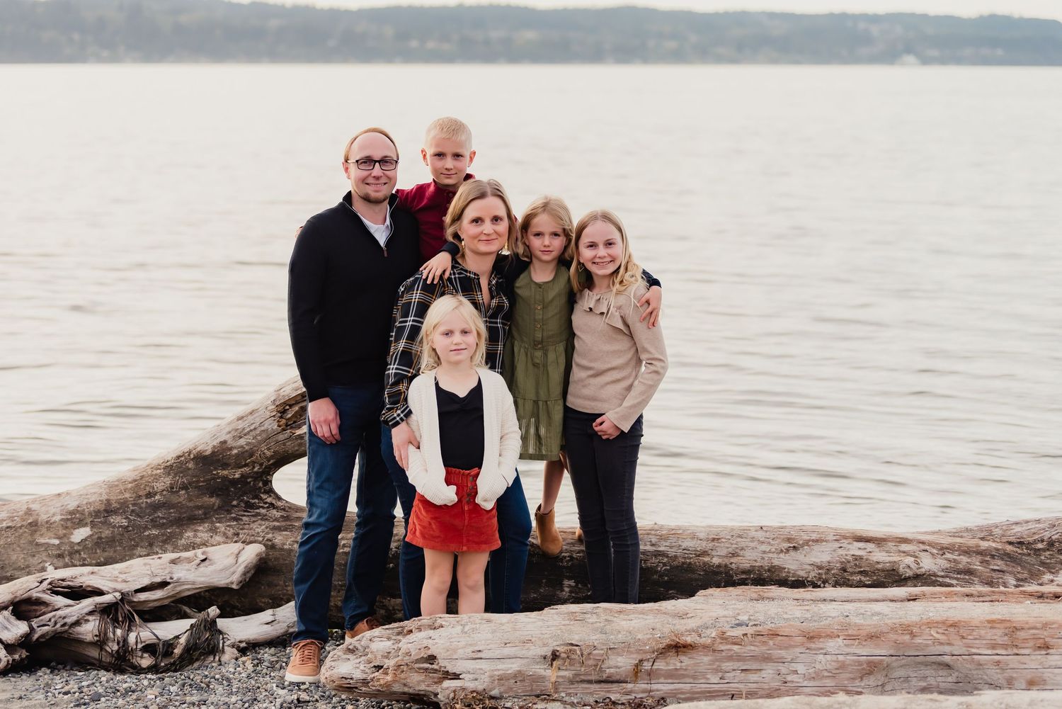 A family sits together on driftwood along a serene waterfront with trees in the background.
