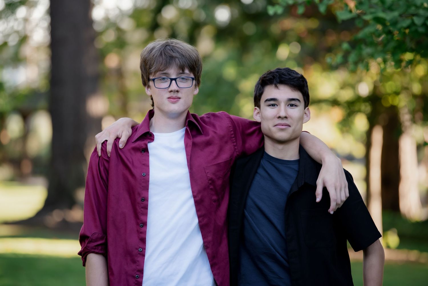 Two young men standing together outdoors, one wearing a burgundy shirt and one in black, with trees in background.