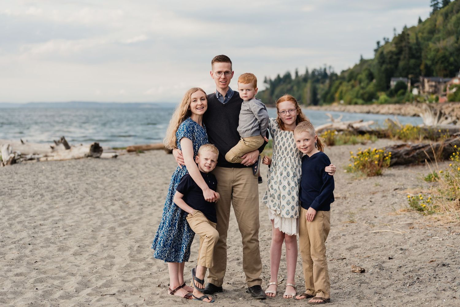 A family wearing coordinated blue and neutral outfits poses together on a Mukilteo, WA beach with driftwood and trees.