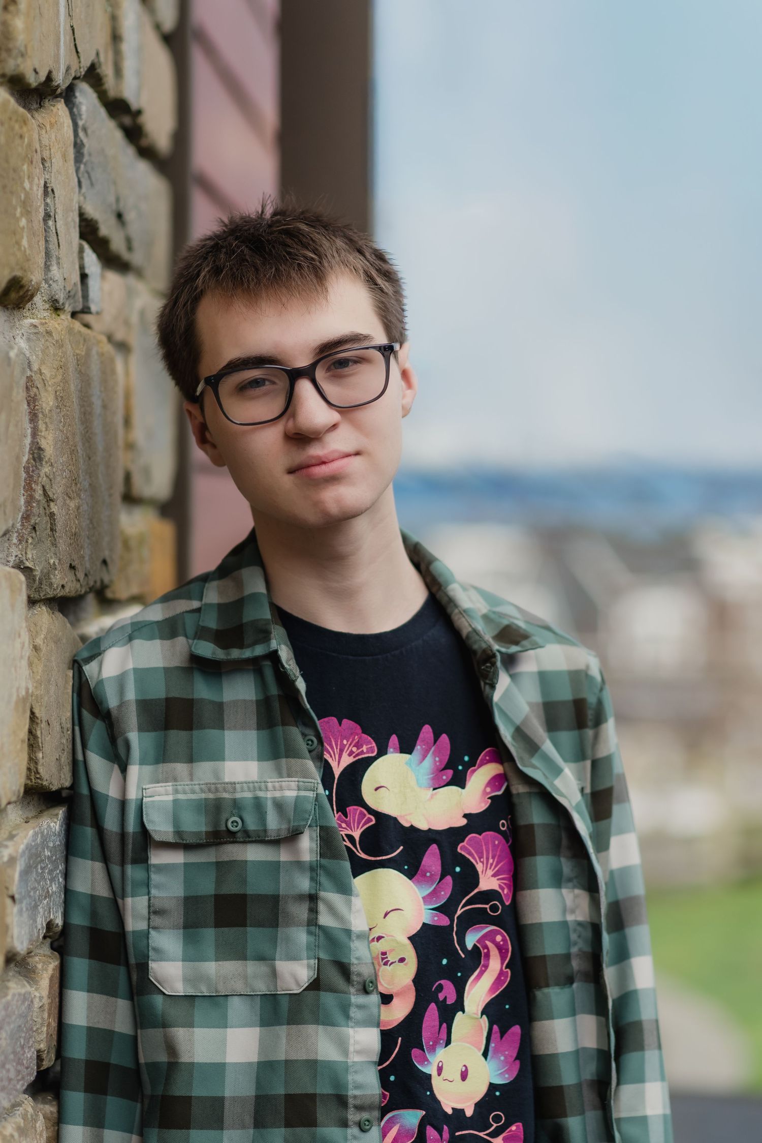 Young man with glasses wearing a plaid flannel shirt over a graphic tee, leaning against a stone wall outdoors.