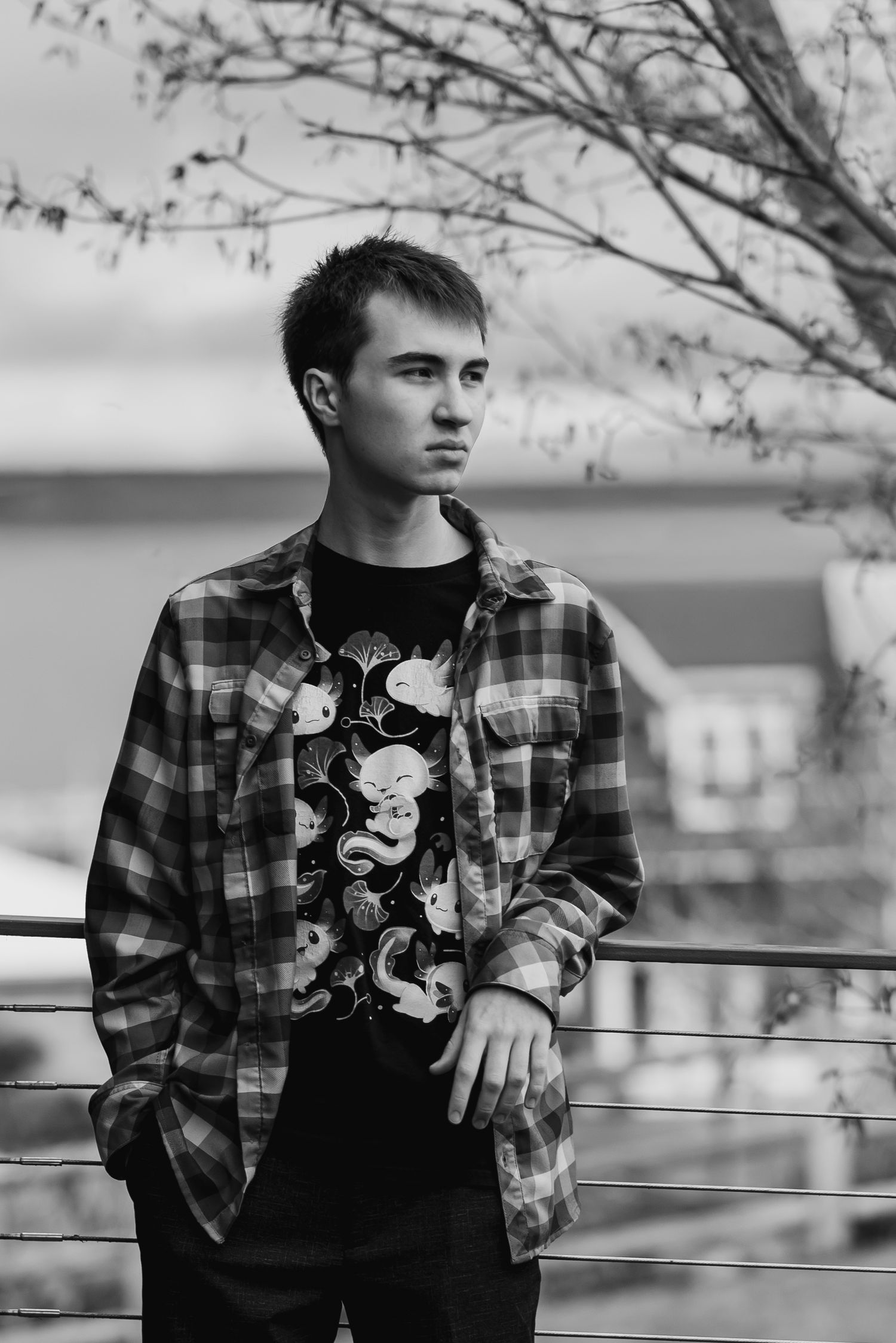 Black and white portrait of a young man in plaid shirt and graphic tee, standing outdoors near a railing.