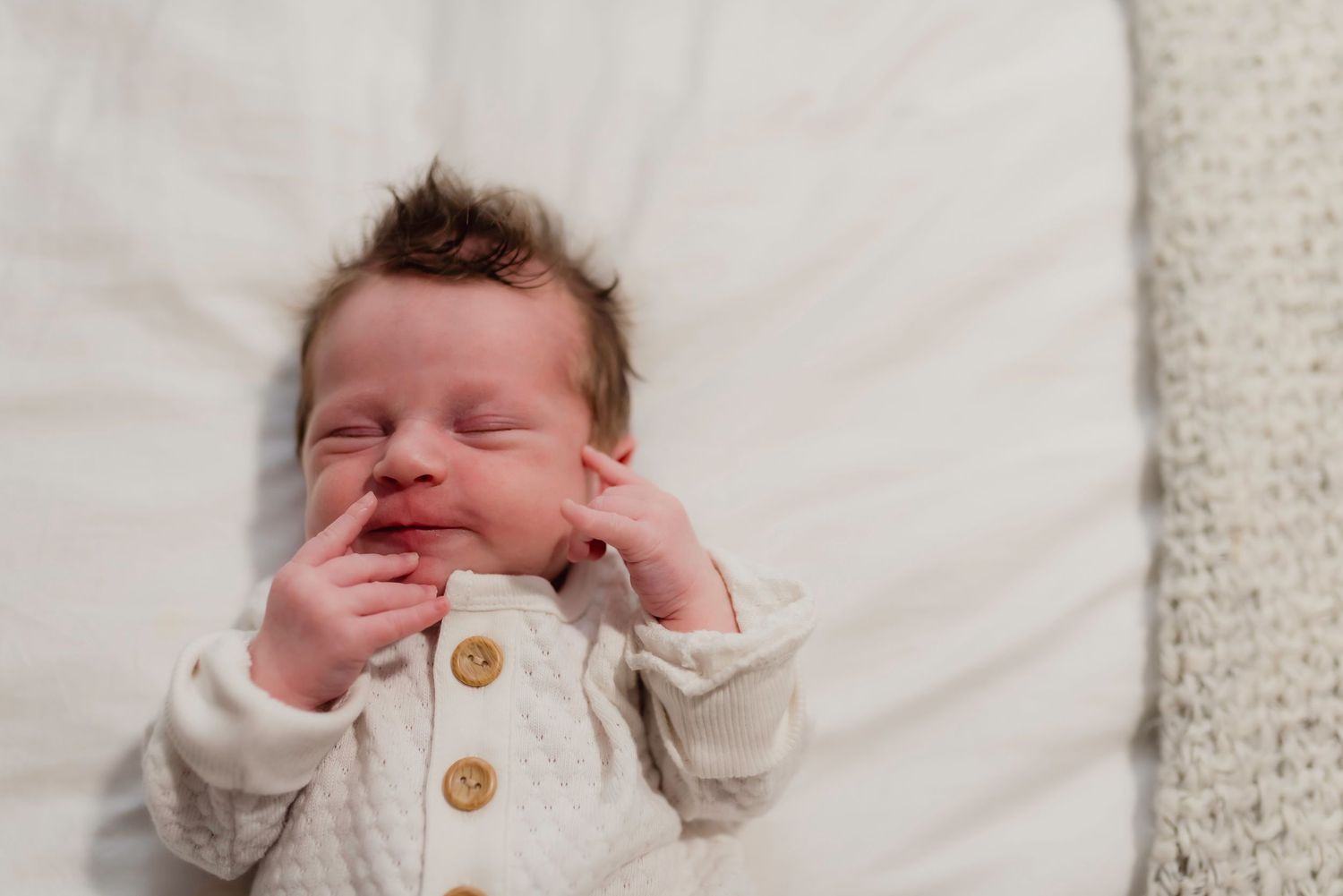 Newborn baby sleeping peacefully in white onesie with wooden buttons, hands near face on soft white blanket in Mukilteo Washington.