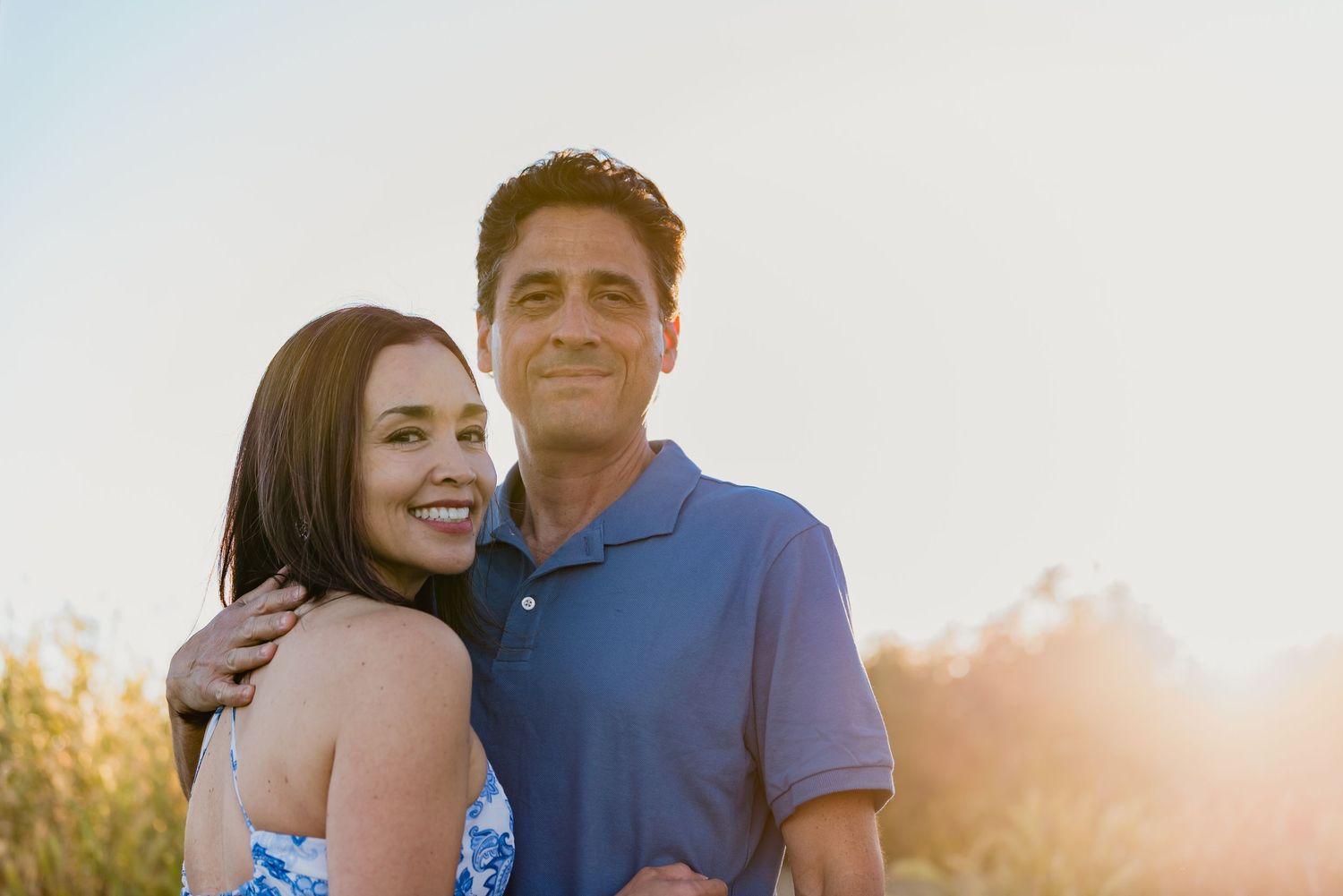 A couple embraces and laughs together during a golden hour photoshoot in a field at sunset.