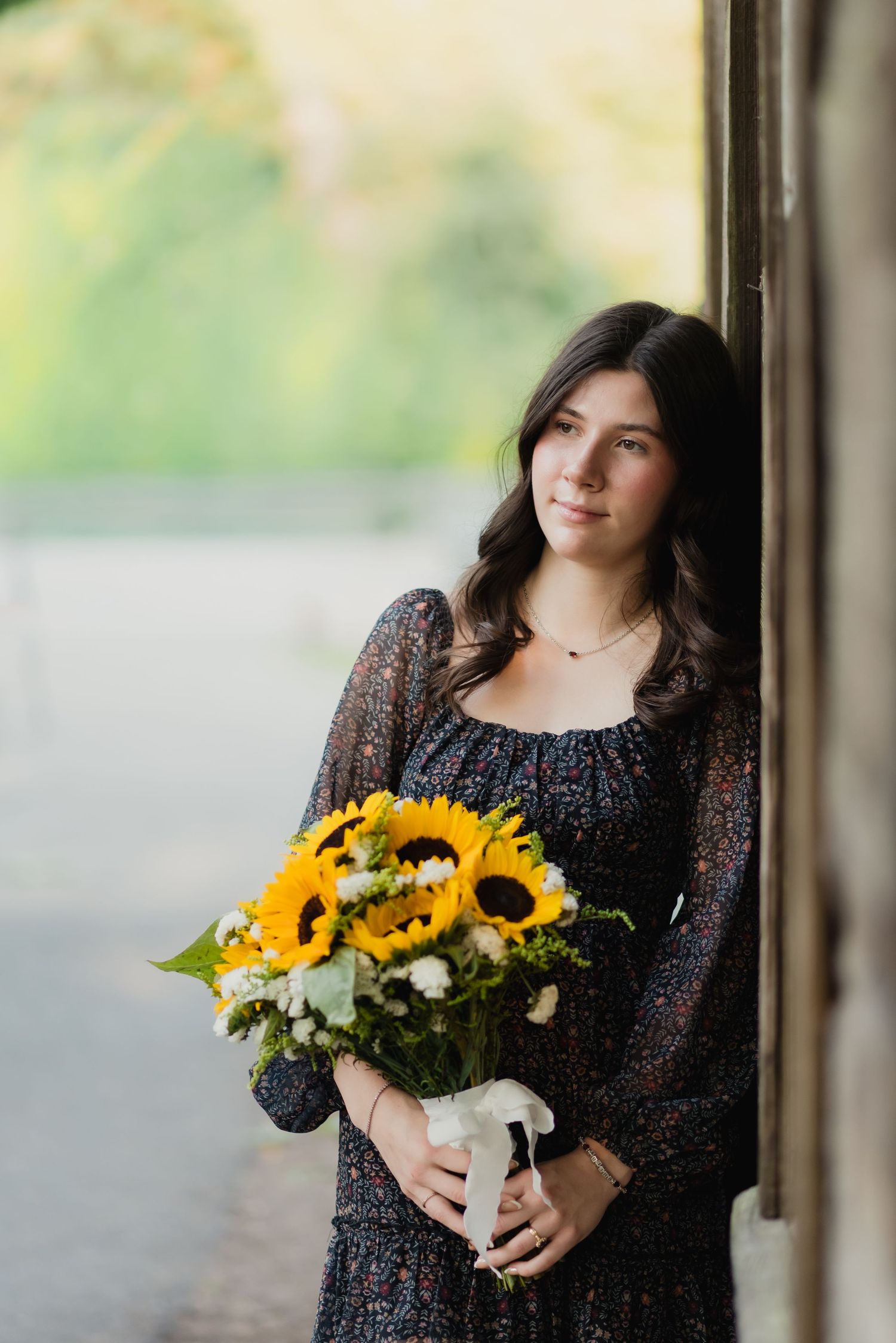 Person in black lace dress holds vibrant sunflowers and white flowers in a vase by a window.